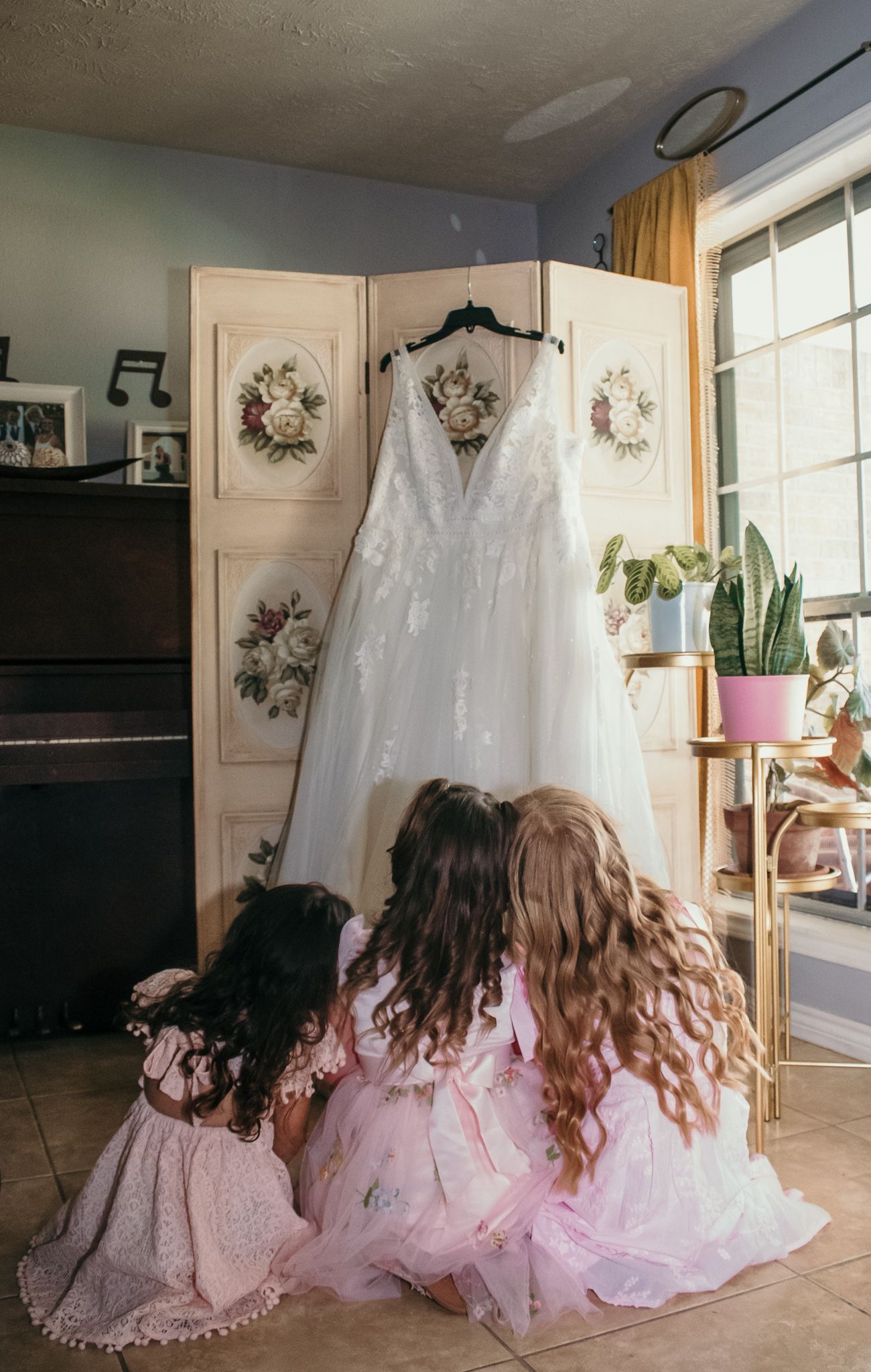 White wedding dress hangs on display as two young attendants in pink dresses admire it.