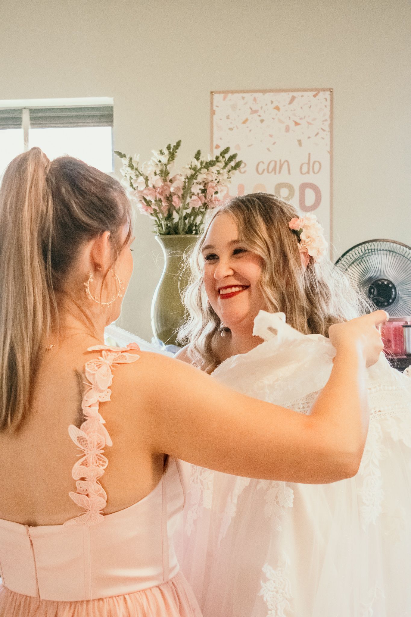 A joyful moment between women in light-colored dresses, with pink flowers and wall art in the background.