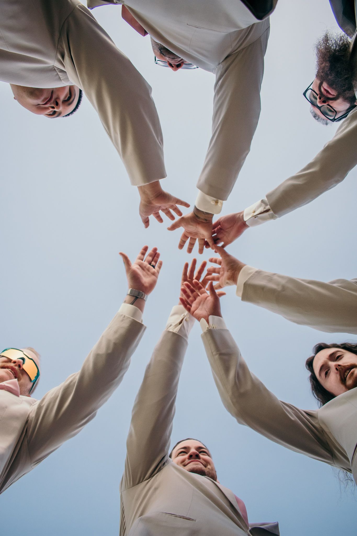 A group of people in cream colored suits join hands together in a circle from a low angle view against the sky.