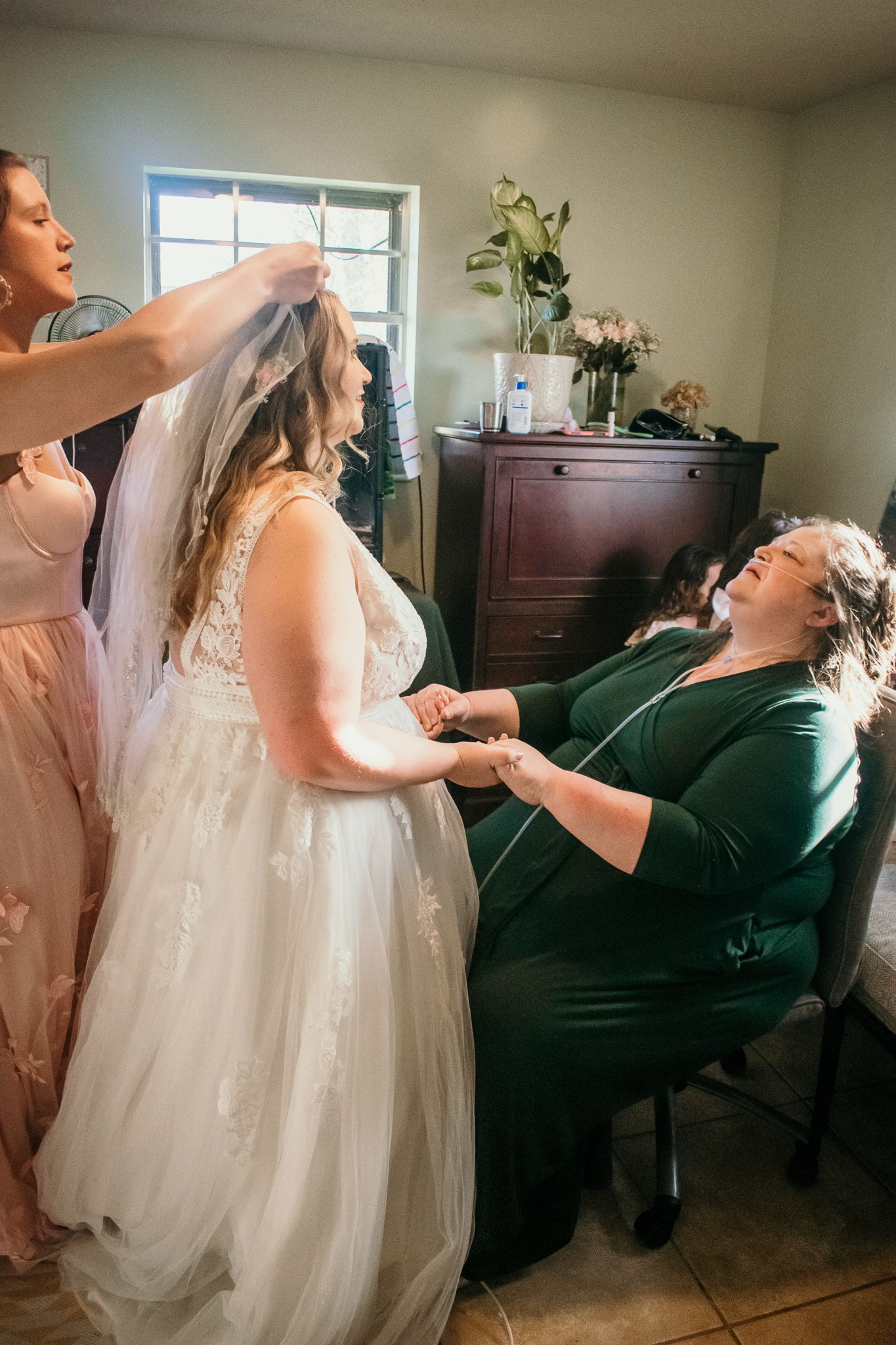 A bride in a flowing white gown stands while getting ready on her wedding day in a room with window light.