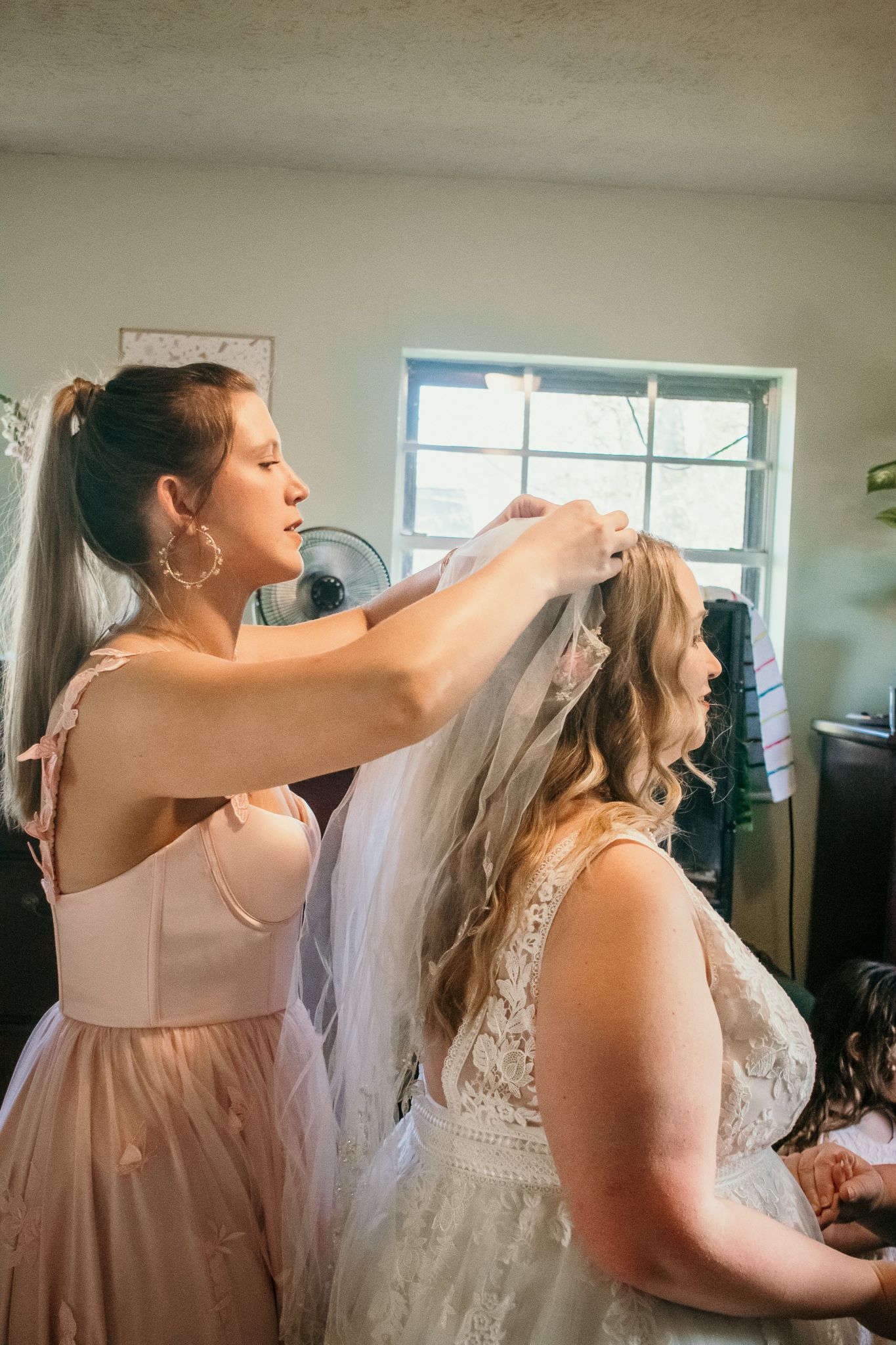 A bridesmaid in a pink dress helps adjust the veil on a bride wearing a lace wedding gown during wedding preparations.