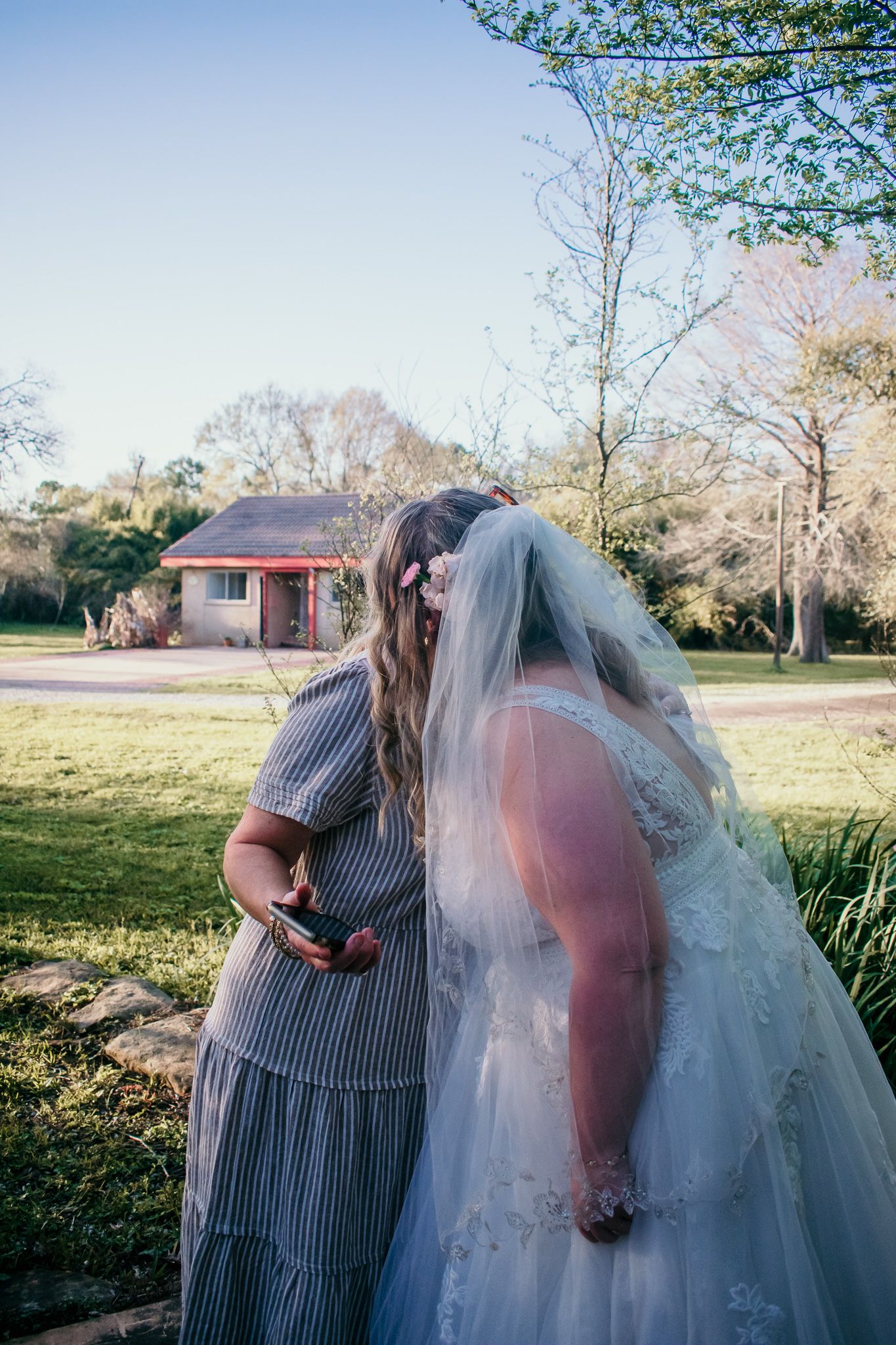 A bride and wedding attendant share an intimate moment outdoors near a small house.