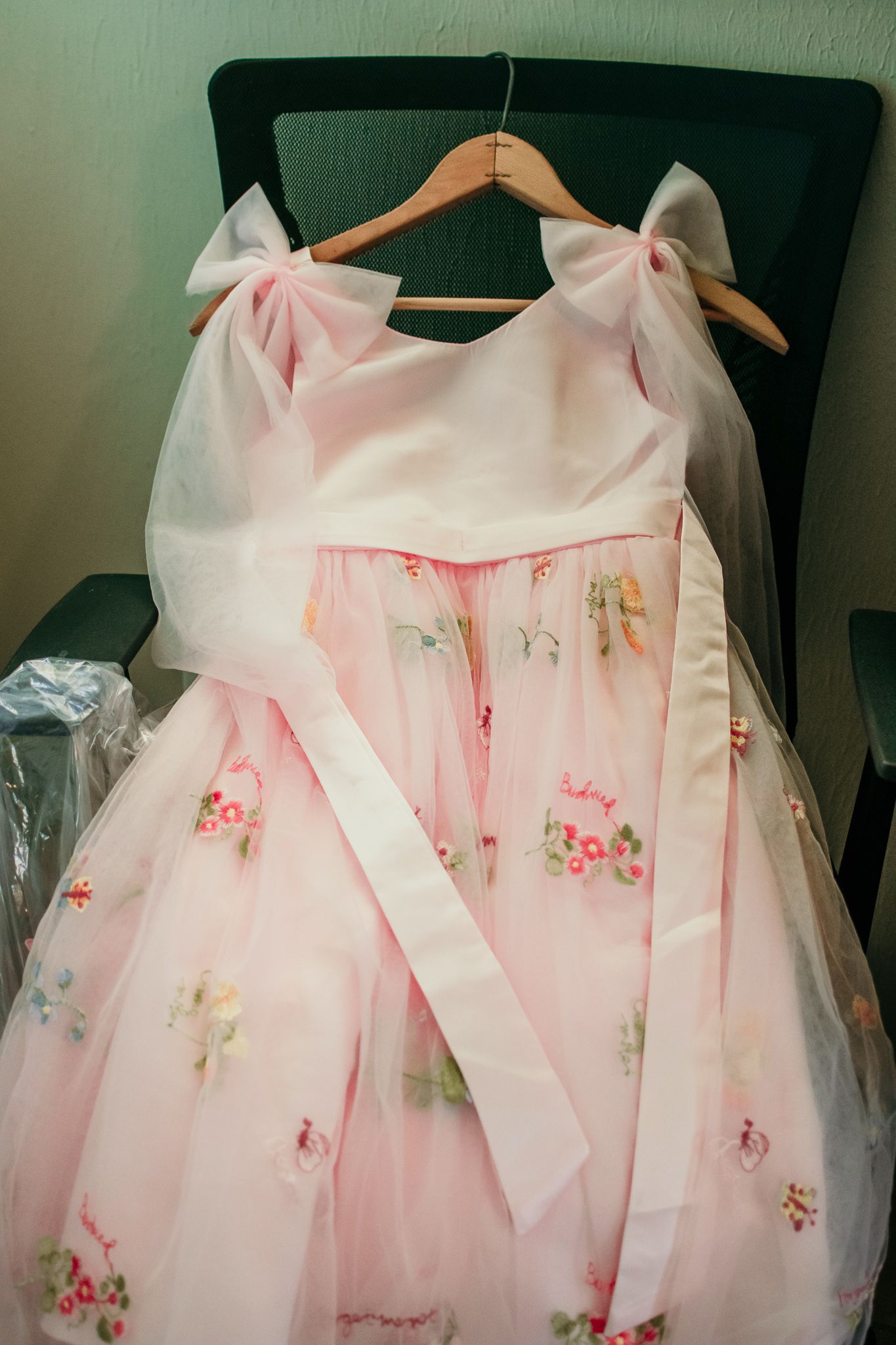 A delicate pink tulle dress with floral embroidery hangs on a wooden hanger against a dark chair.
