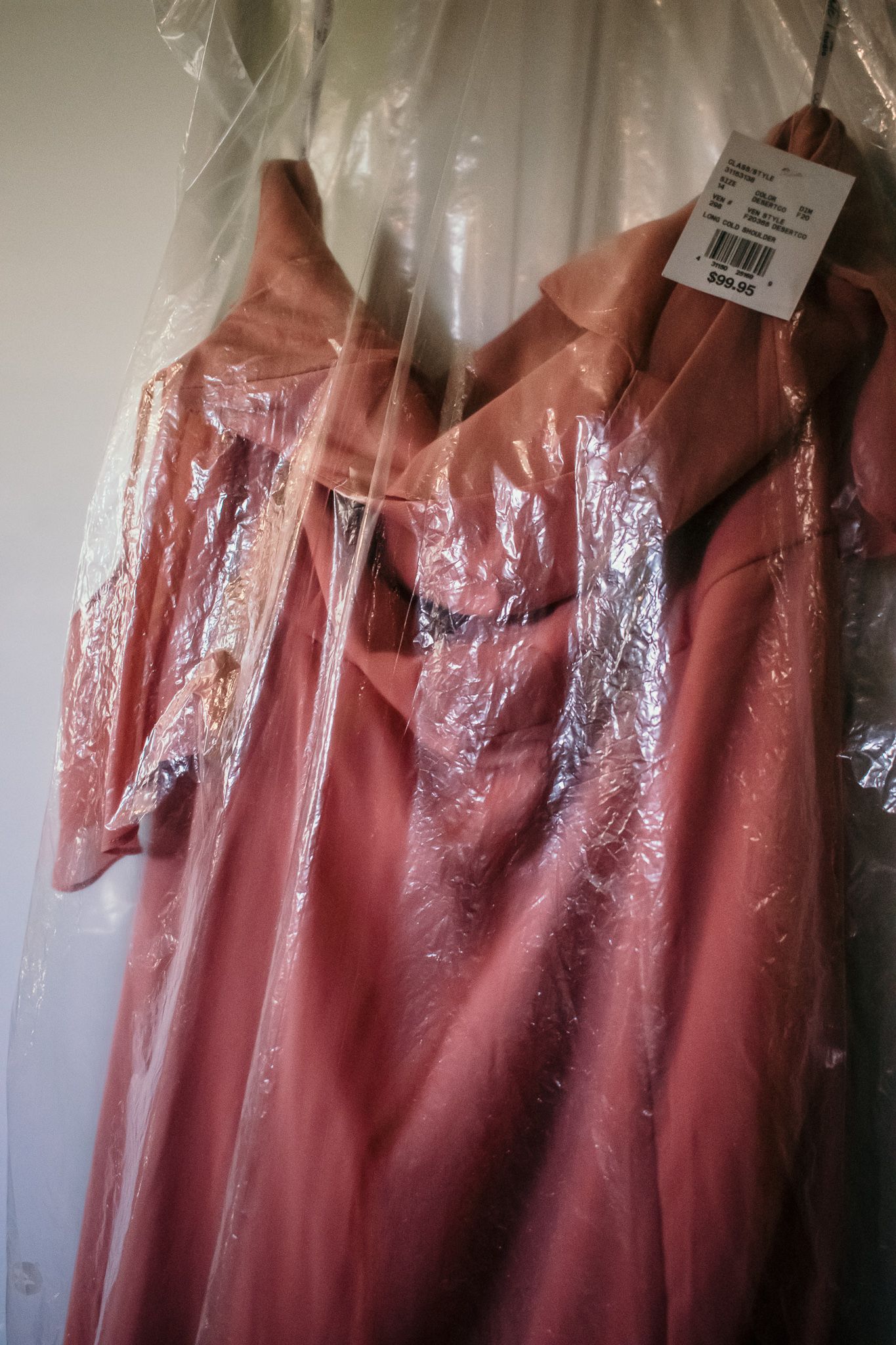 A pink bridesmaid dress hanging in clear plastic garment bag with price tag visible.