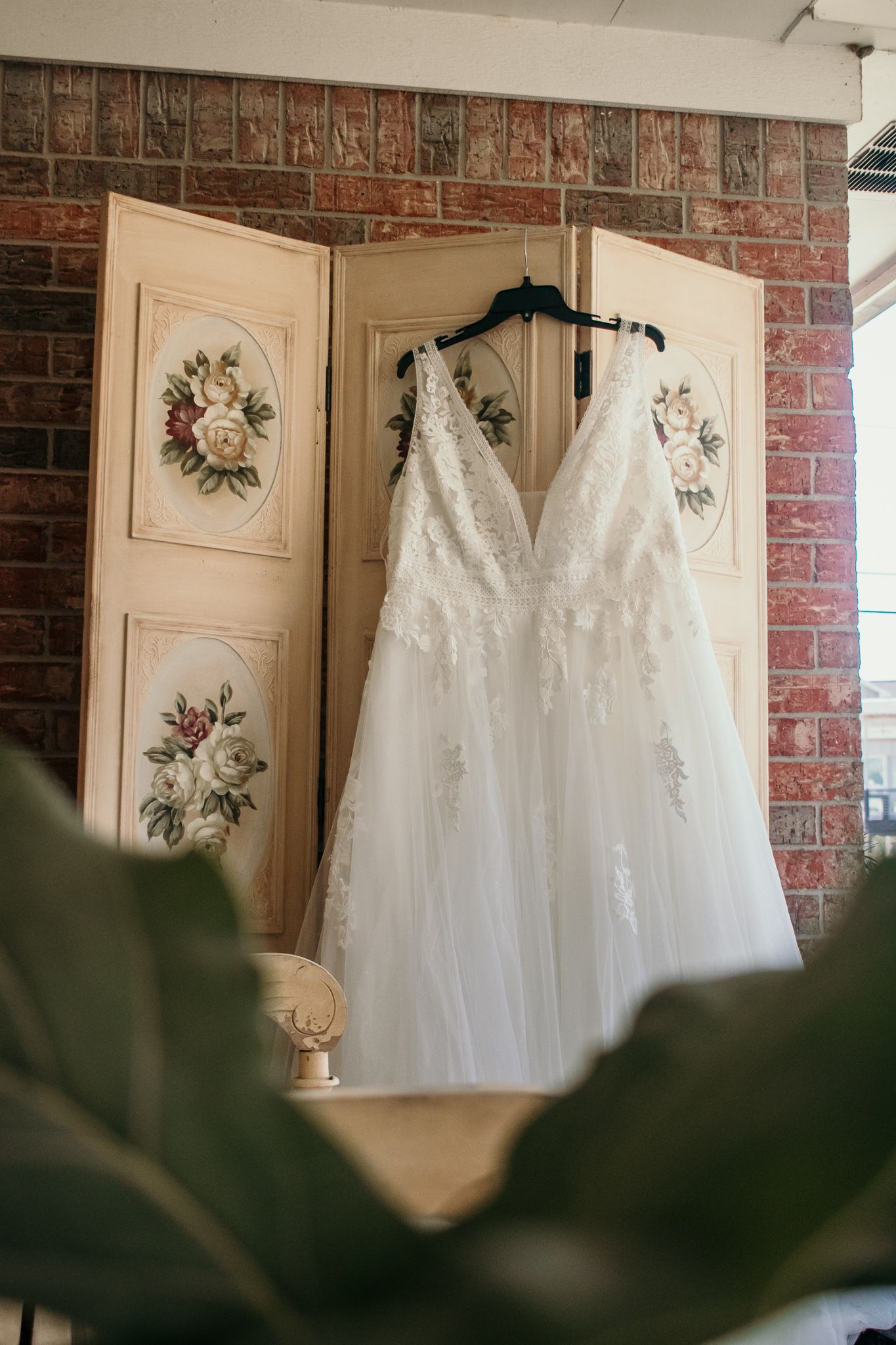 Elegant white wedding dress displayed on vintage floral folding screen against brick wall.