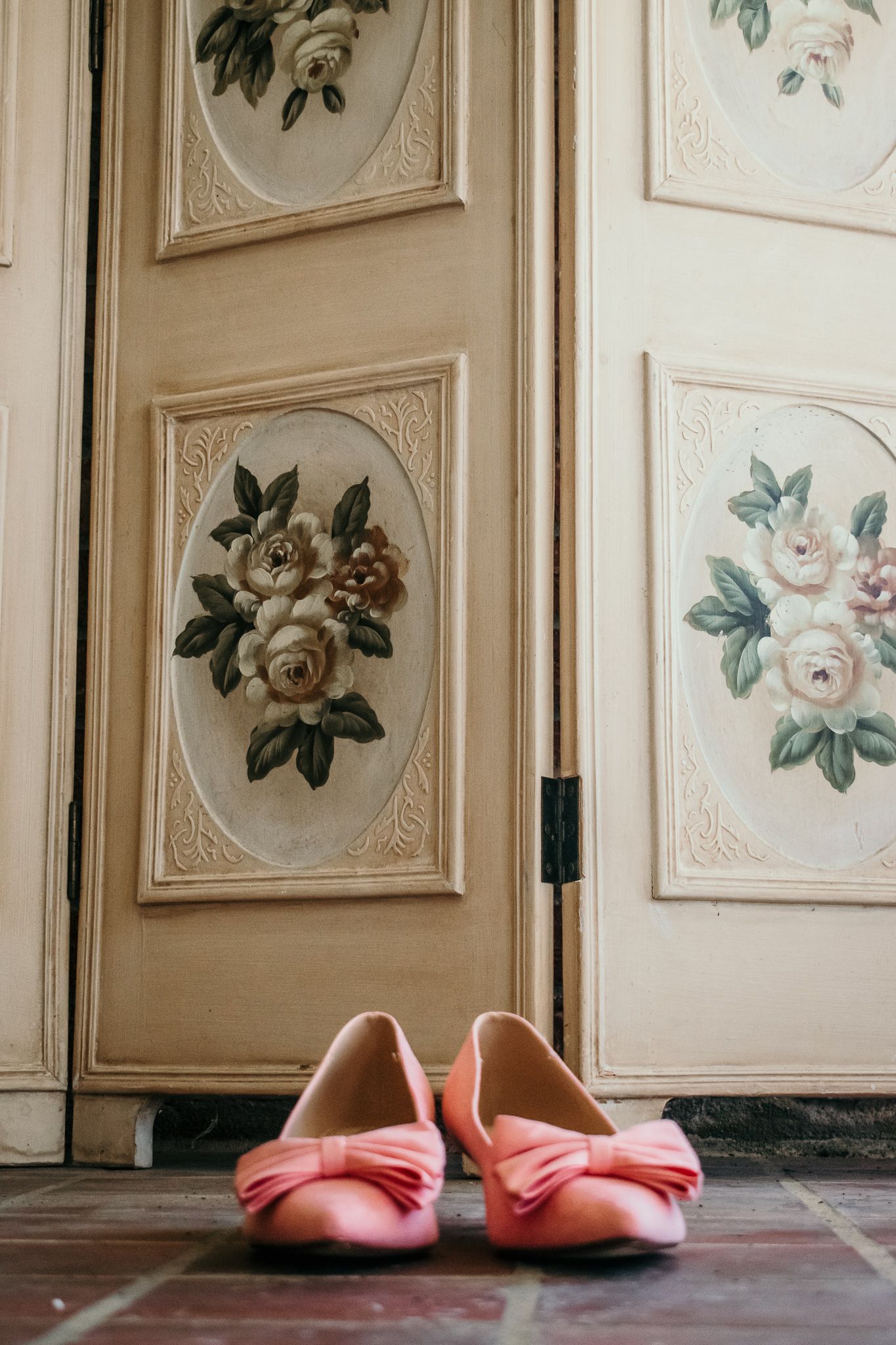 Pink ballet flats sit on a wooden floor against an ornate cabinet decorated with vintage floral paintings.