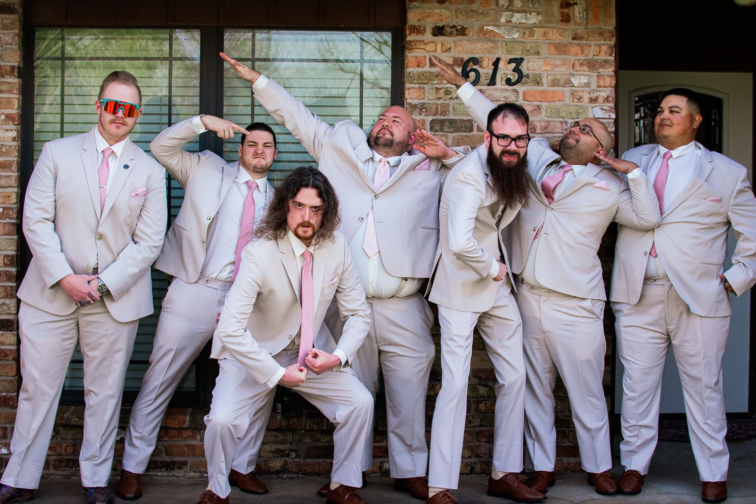 Groomsmen in light colored suits strike playful poses against a brick wall.