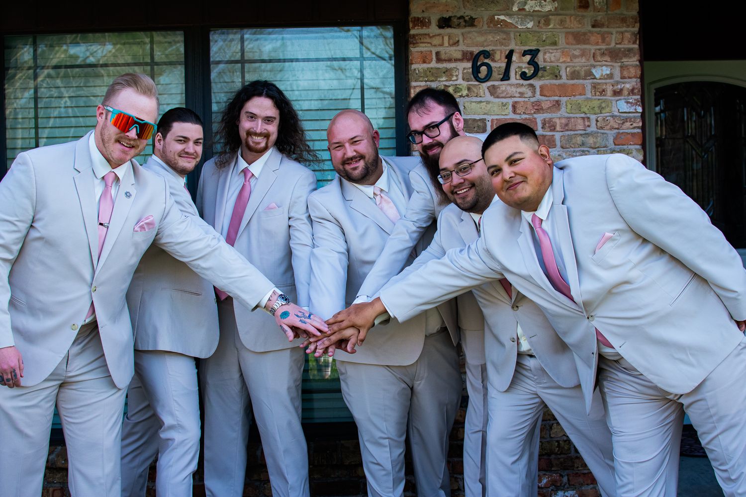 Groomsmen in matching light gray suits and pink ties pose playfully together against a brick wall.