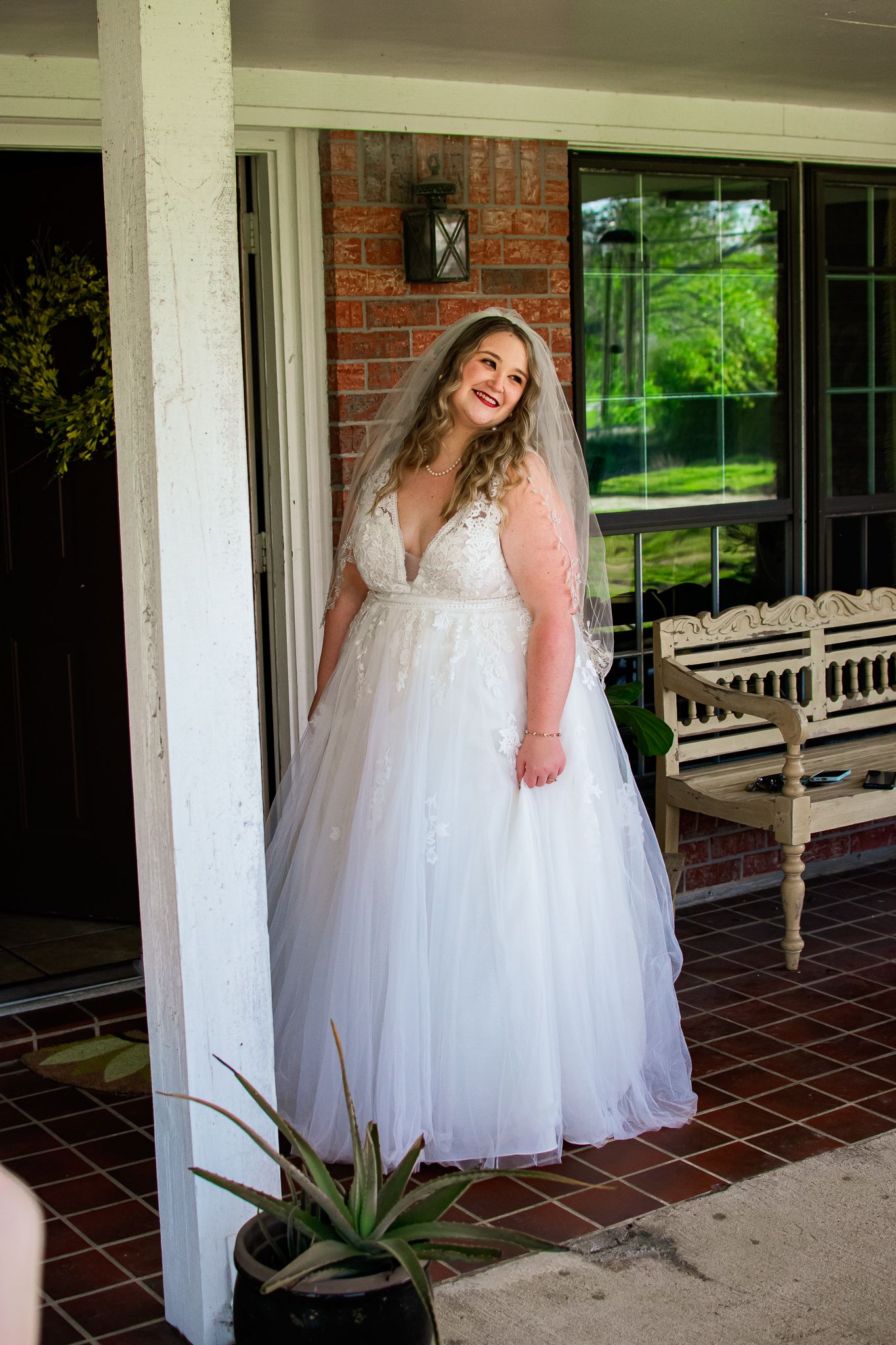 Beautiful bride in flowing white wedding dress smiles on a brick porch with a wooden bench and potted plant.