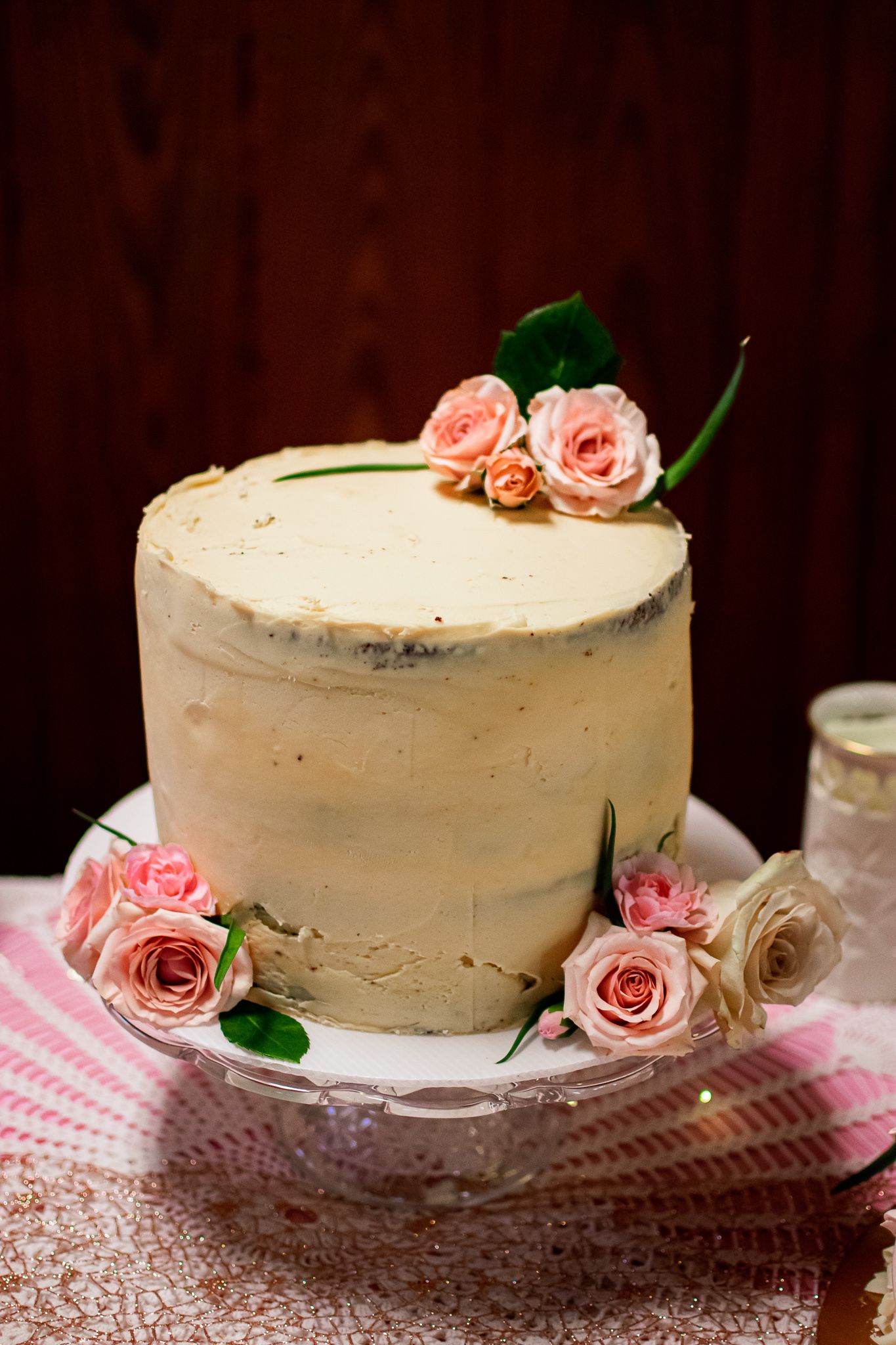 A rustic white frosted cake decorated with delicate pink roses on a pink checkered tablecloth against a dark background.