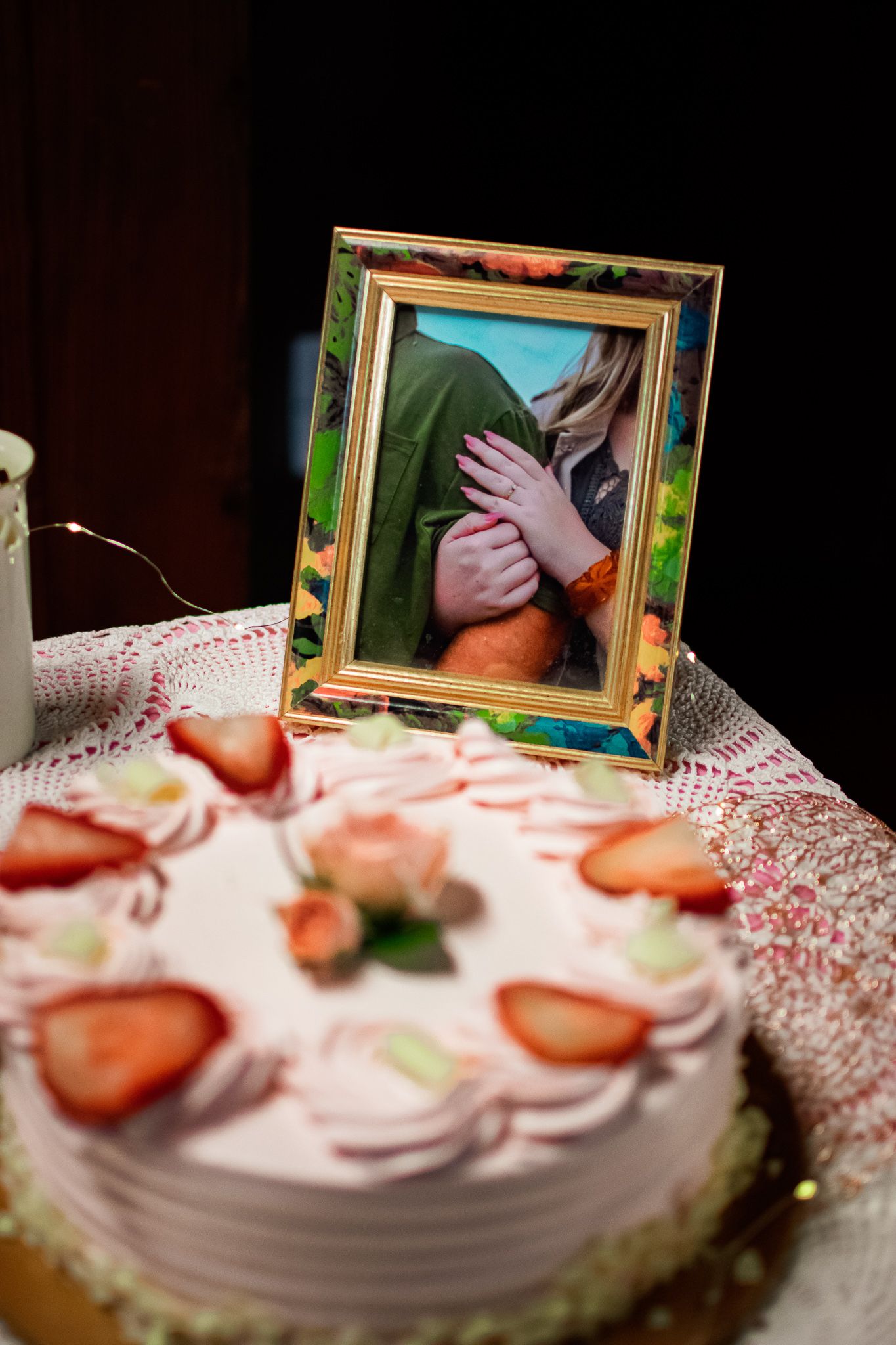 A pink strawberry cake with whipped cream frosting sits beside a decorative framed photograph on a lace tablecloth.