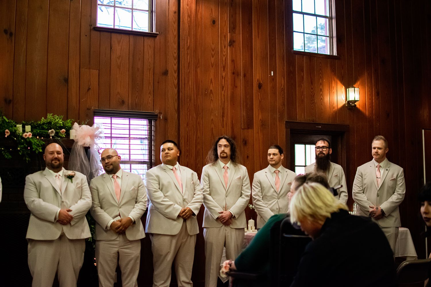 Wedding party in light colored suits standing in line inside a rustic wooden chapel.