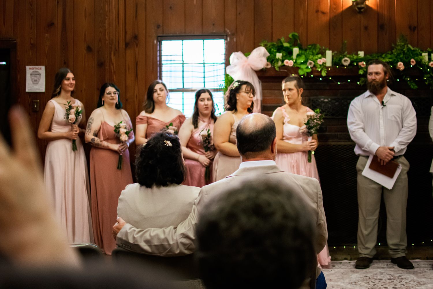 Wedding ceremony scene with bridesmaids in pink dresses standing in front of wood paneled wall with greenery.