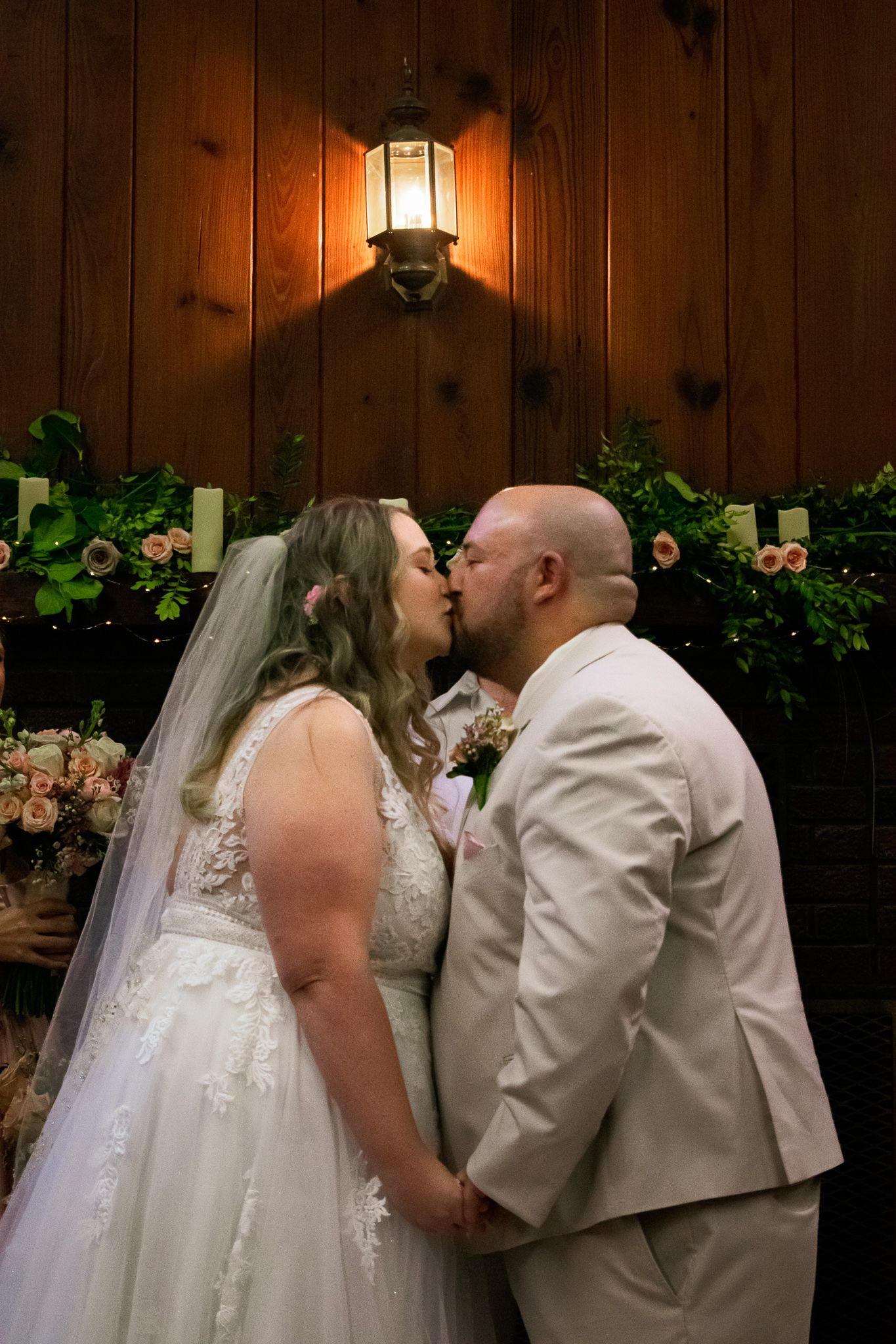 A romantic wedding kiss moment beside a glowing lantern on a wooden wall with floral decorations.