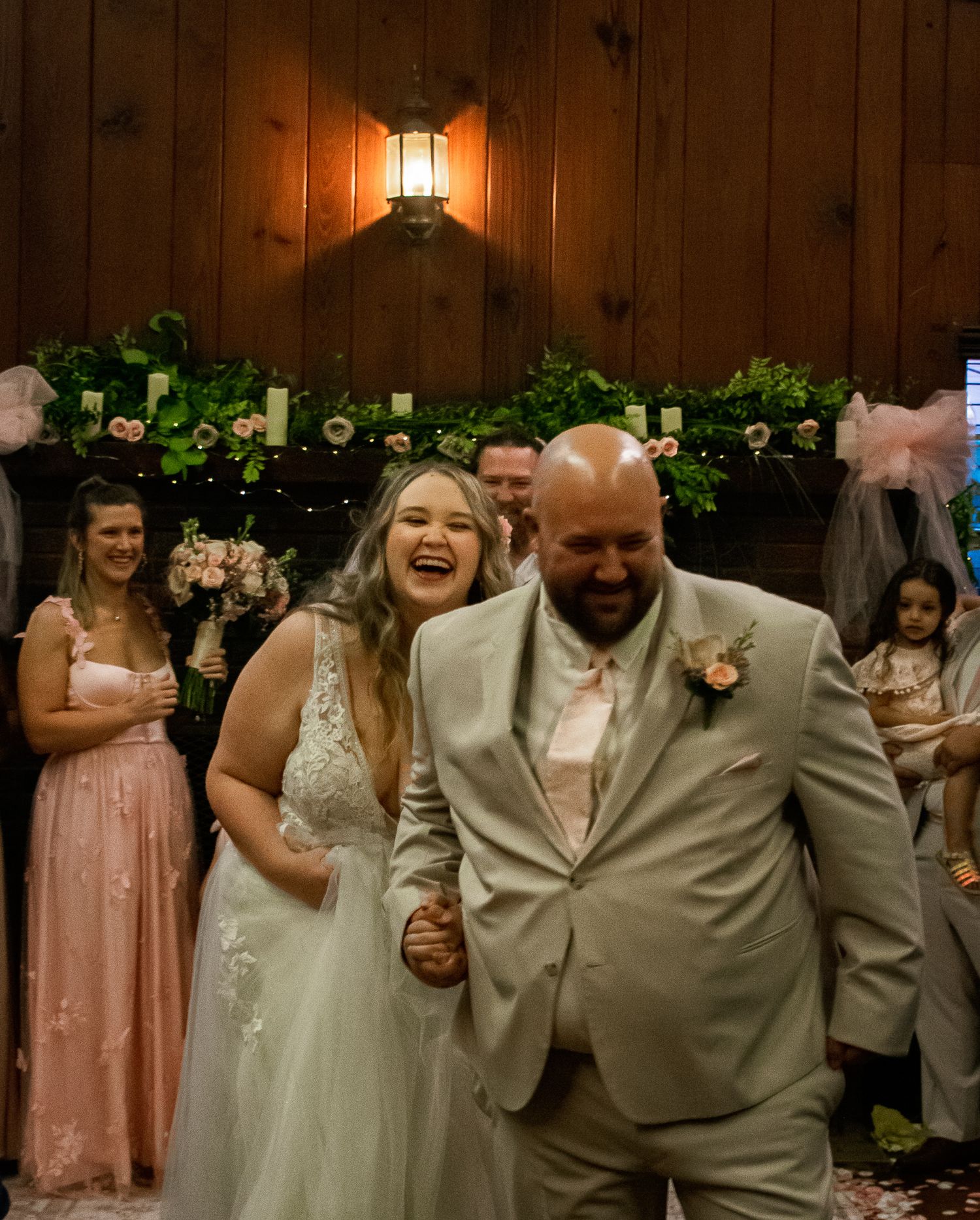 A joyful wedding couple laughing while walking down the aisle decorated with greenery and lights.
