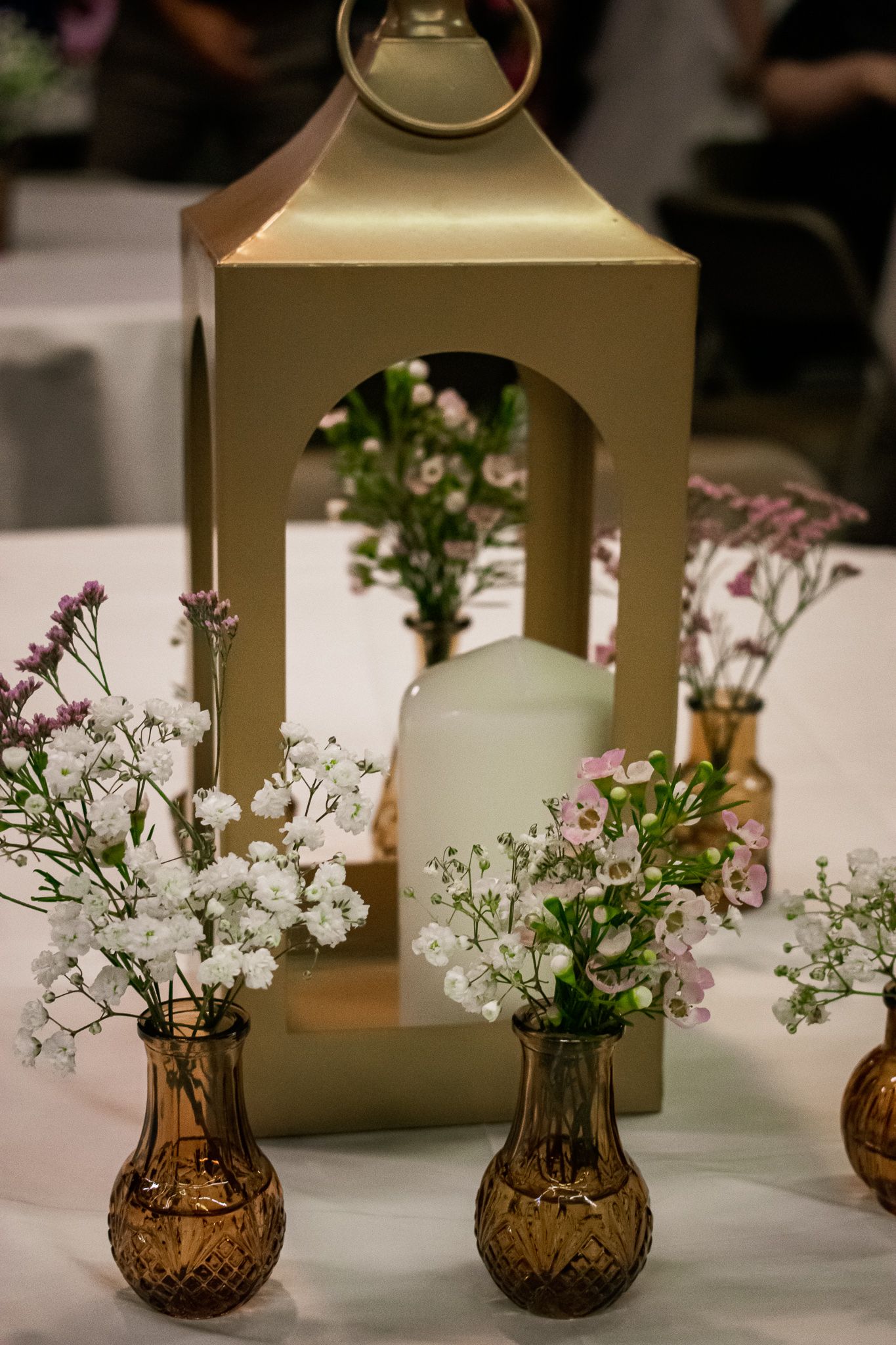 Gold lantern centerpiece with delicate baby's breath flowers in amber glass vases on white tablecloth.