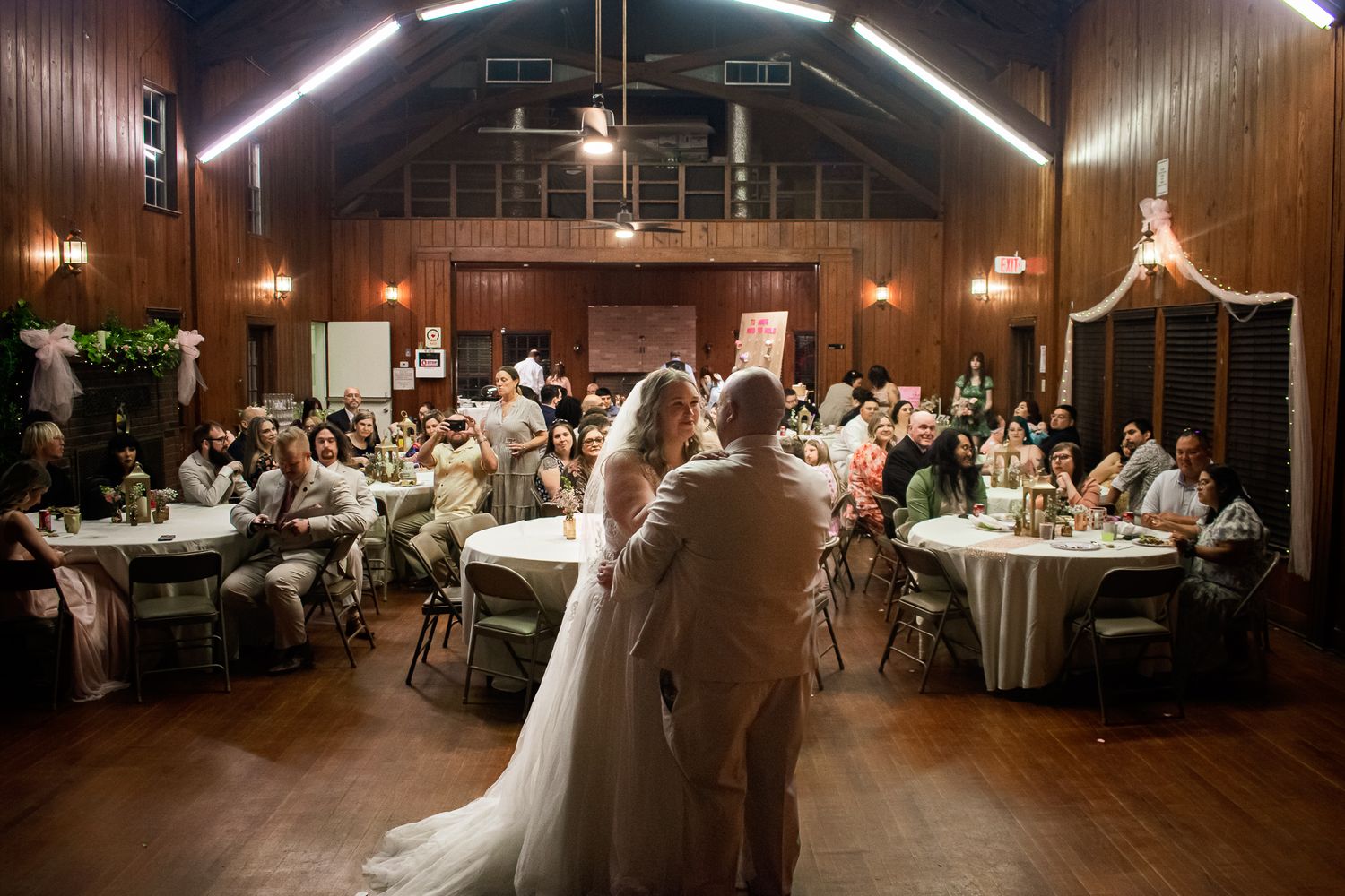 Wedding reception guests seated at round tables inside a rustic wooden barn venue with overhead string lights.