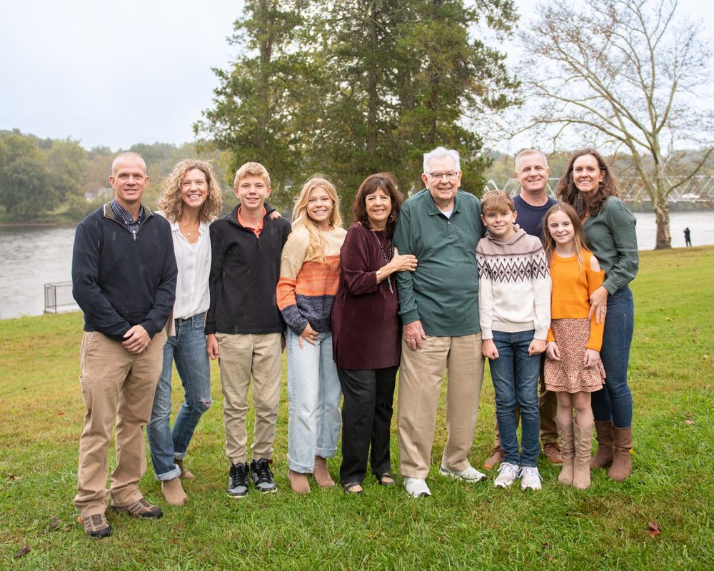 Family group standing together on grass near a lake on an overcast autumn day.