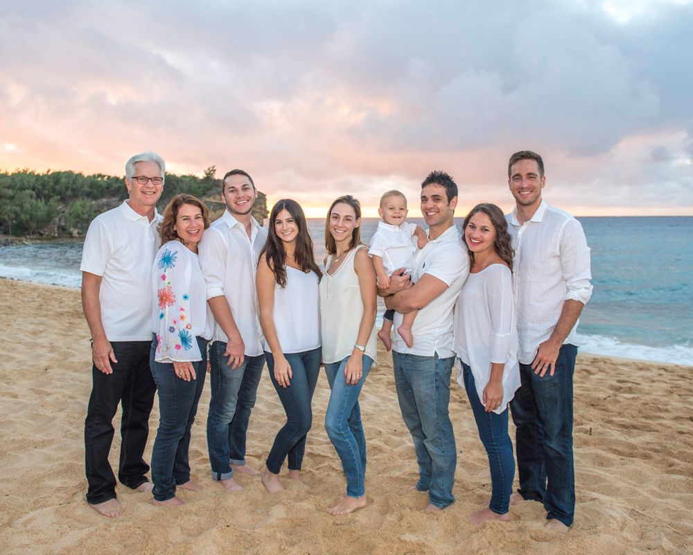 Family group in white and blue clothing poses together on beach at sunset.