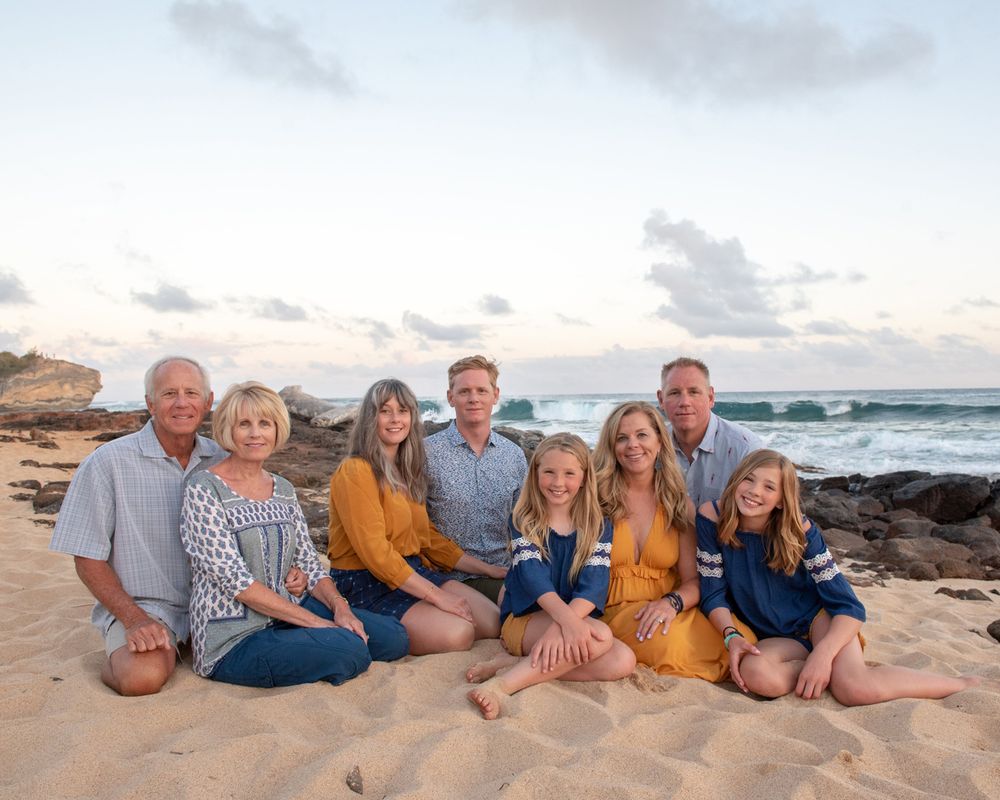 Group sitting together on sandy beach with ocean waves and rocks in background.