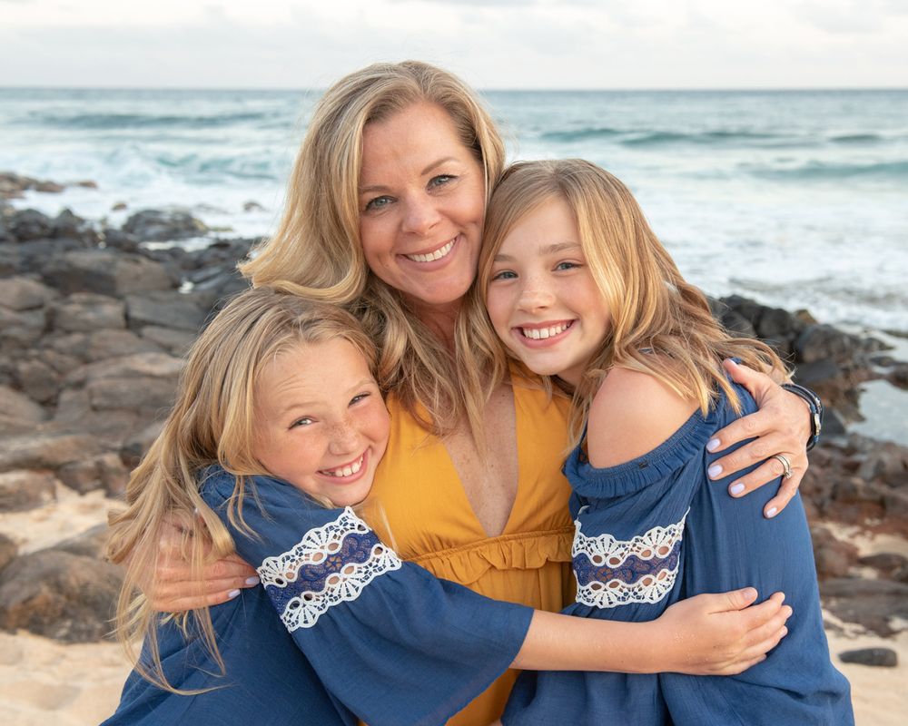 Three people share laughs and hugs on a rocky beach with ocean waves in the background.