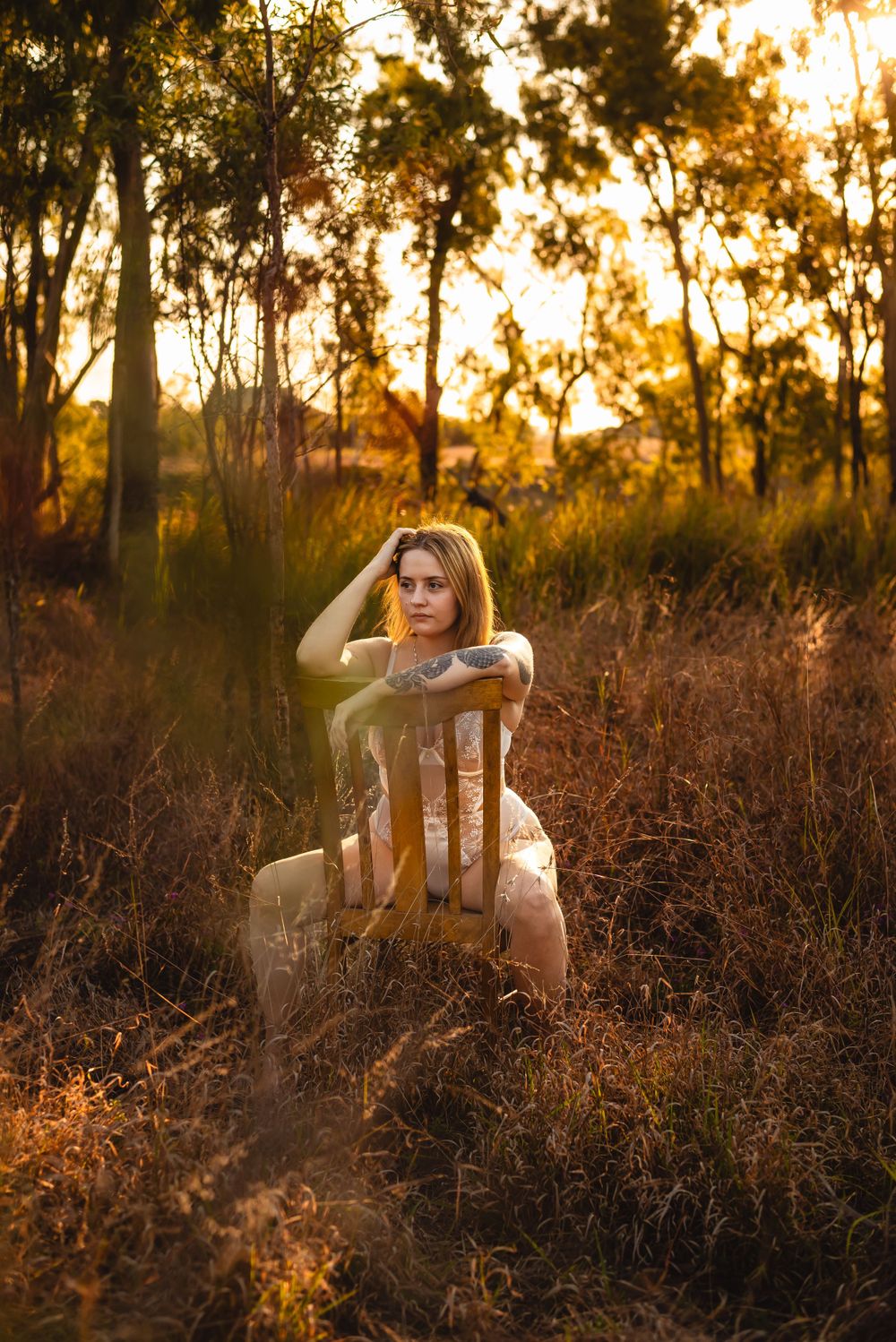 Artistic portrait with model posed on wooden chair in golden hour field.