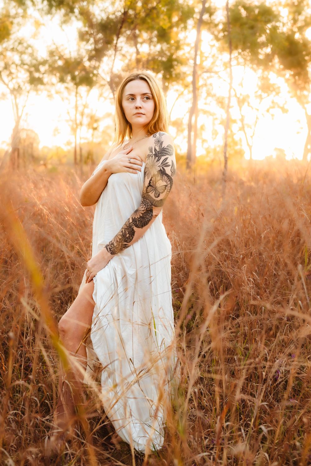 Woman in flowing white dress stands in tall grass at sunset with detailed arm tattoo.