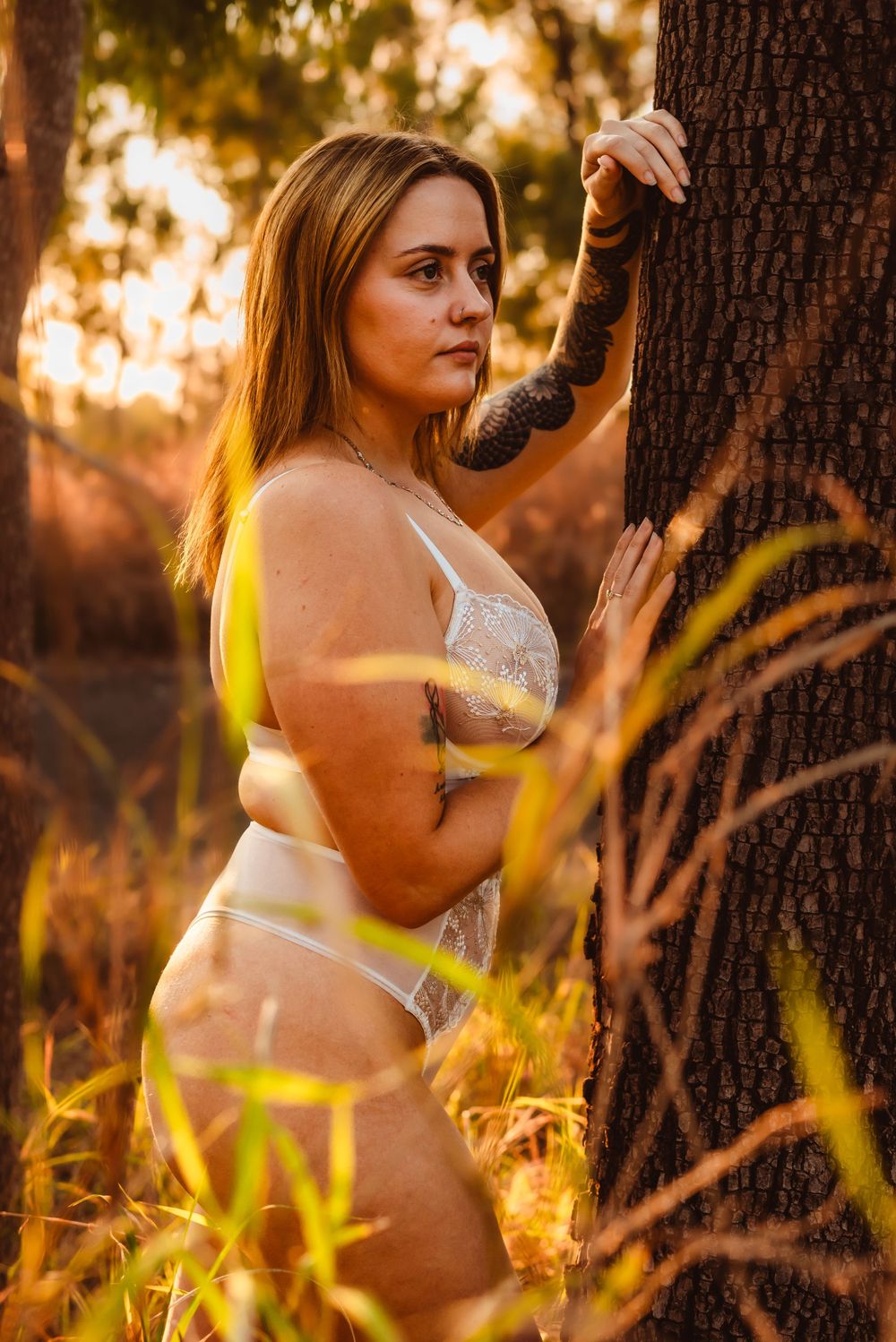 Model in white lingerie poses against tree trunk in golden sunset light.