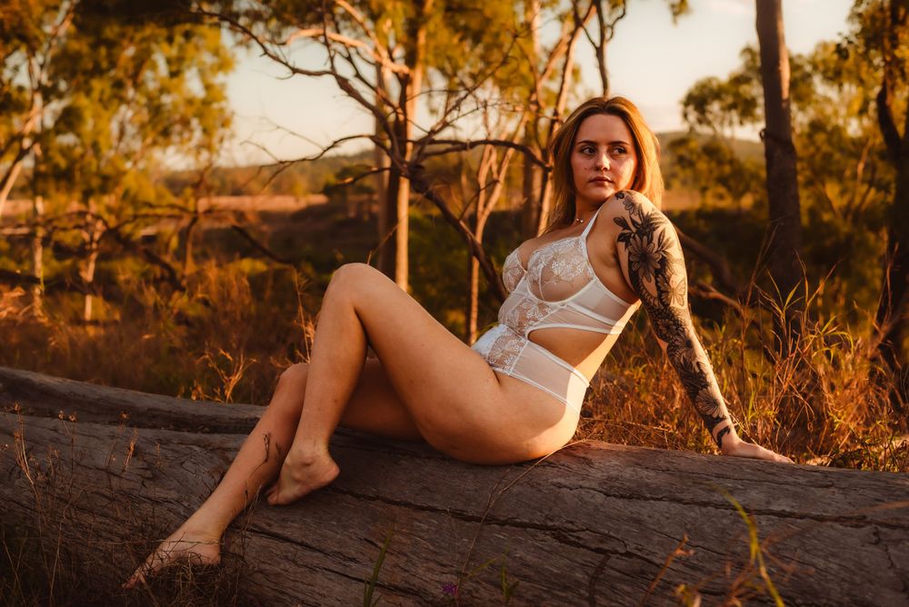 Model reclines on fallen log in white bodysuit during sunset in nature.