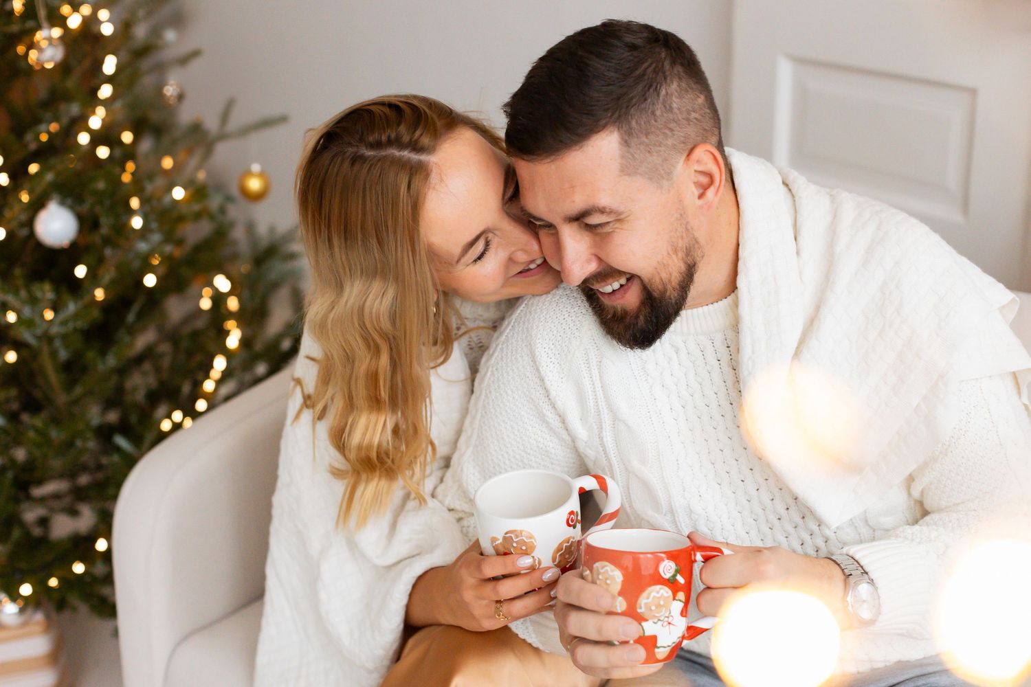 Happy couple sharing a romantic moment by a Christmas tree while holding festive red mugs.