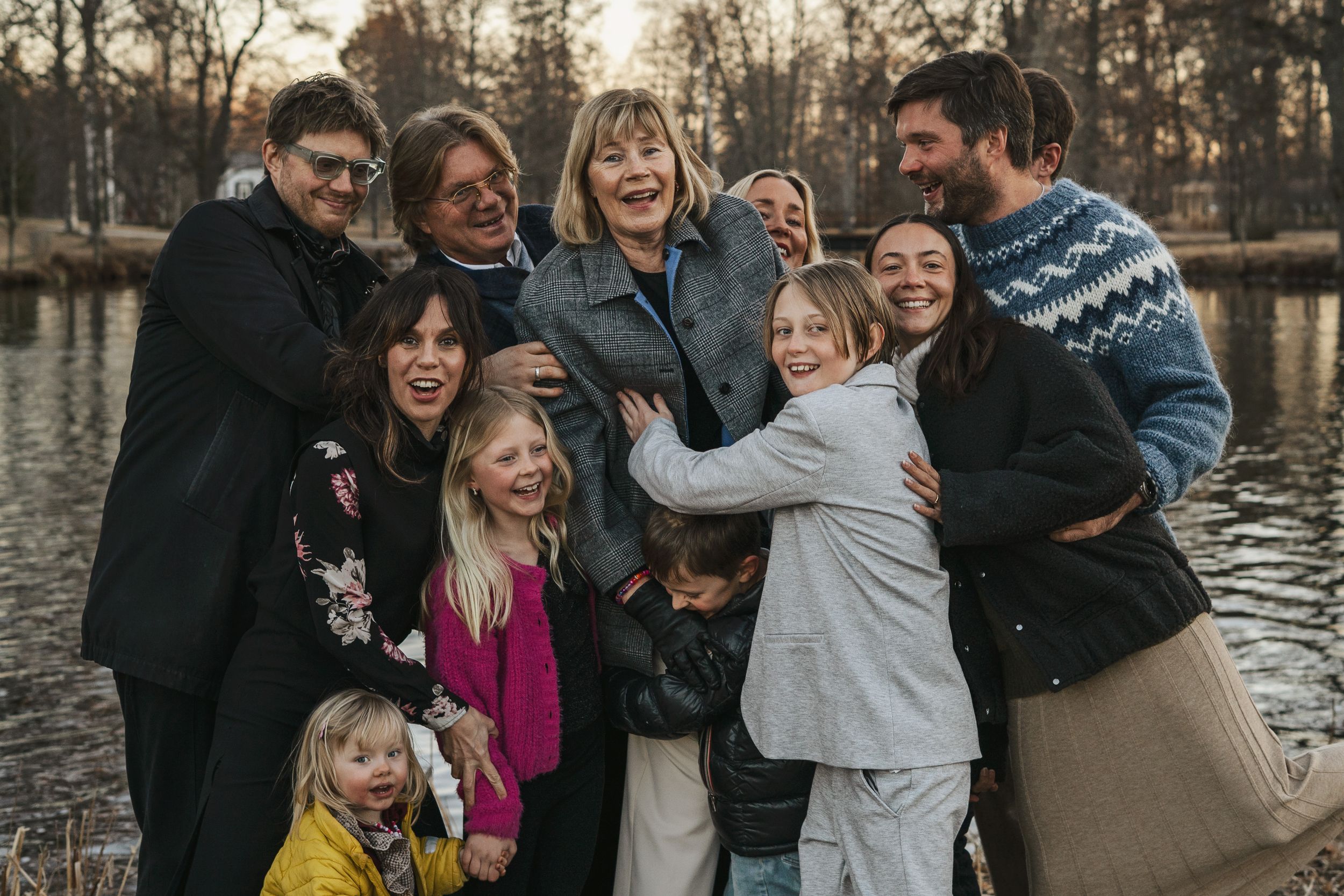 En stor familj skrattar och omfamnar varandra medan de står nära en sjö under solnedgången i vinterjackor. -Fotograf Emma Matkoski