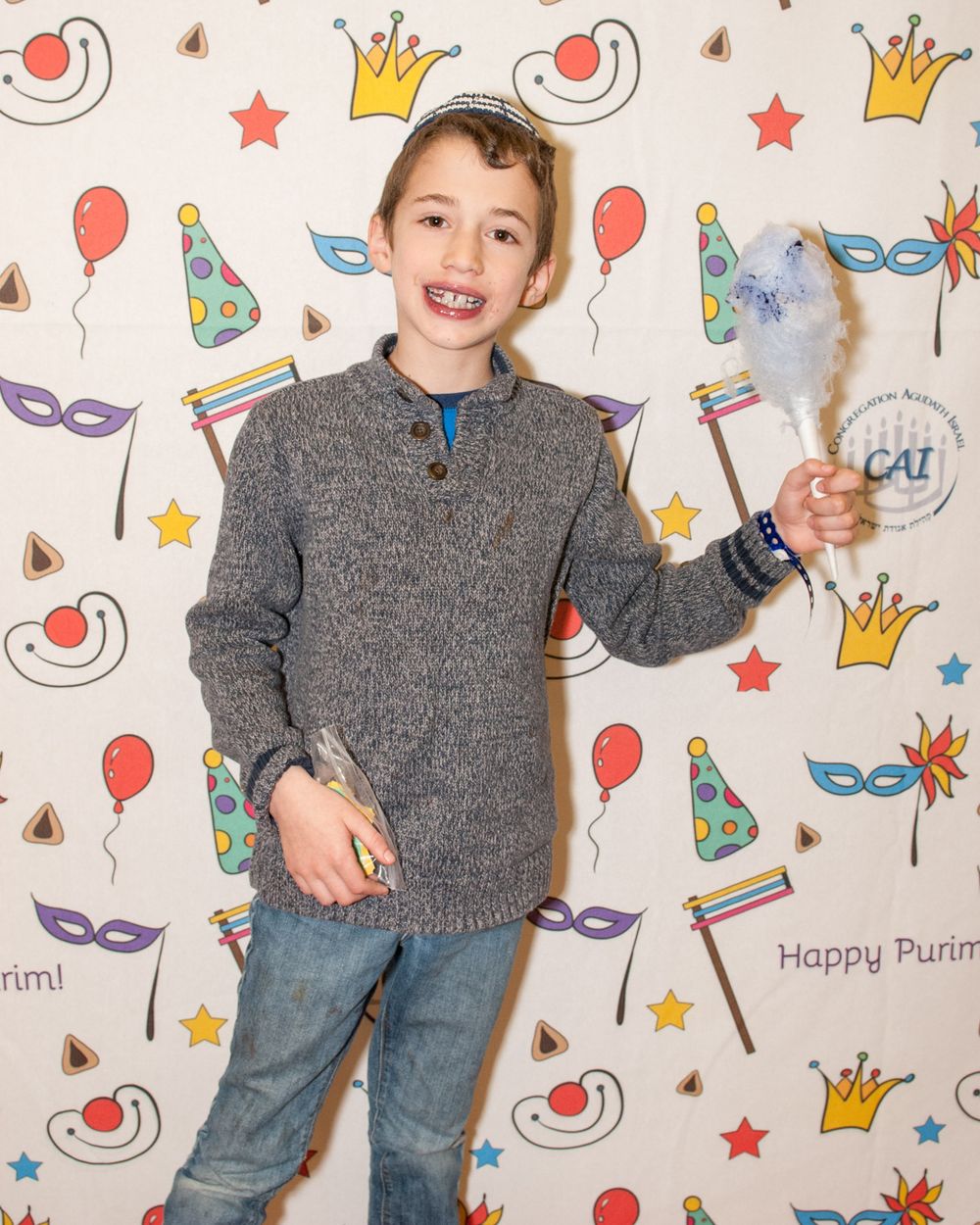 Child in grey sweater poses with cotton candy against festive party backdrop with crown patterns.