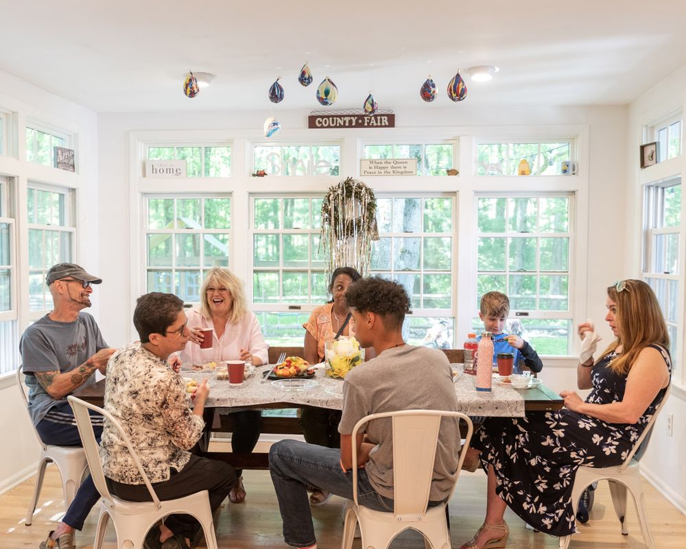 A group gathers around a dining table in a bright sunlit room with large windows.