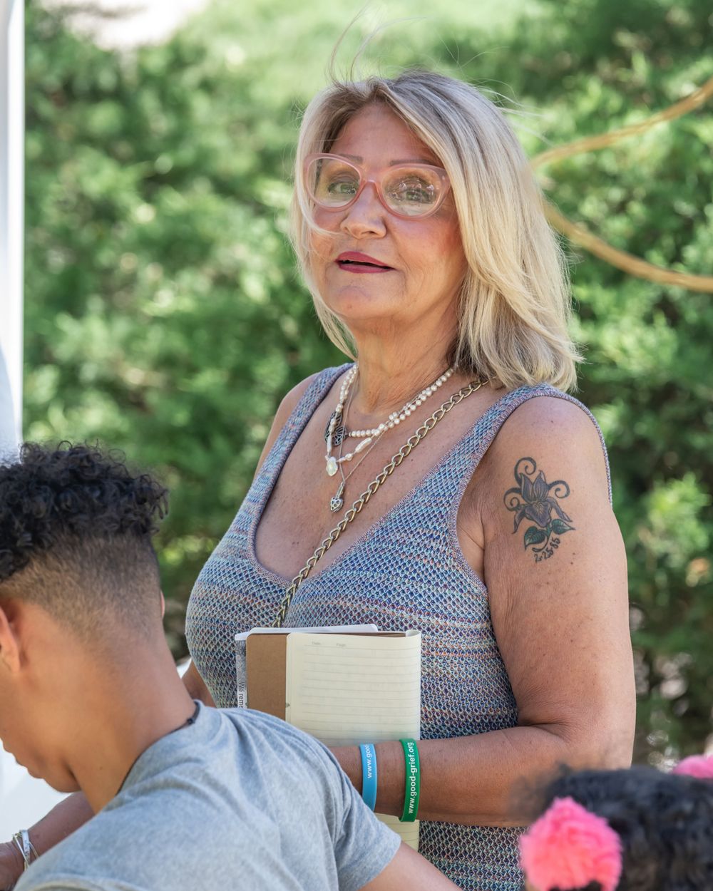 A person in a gray tank top with tattoos and jewelry stands outdoors in summer.