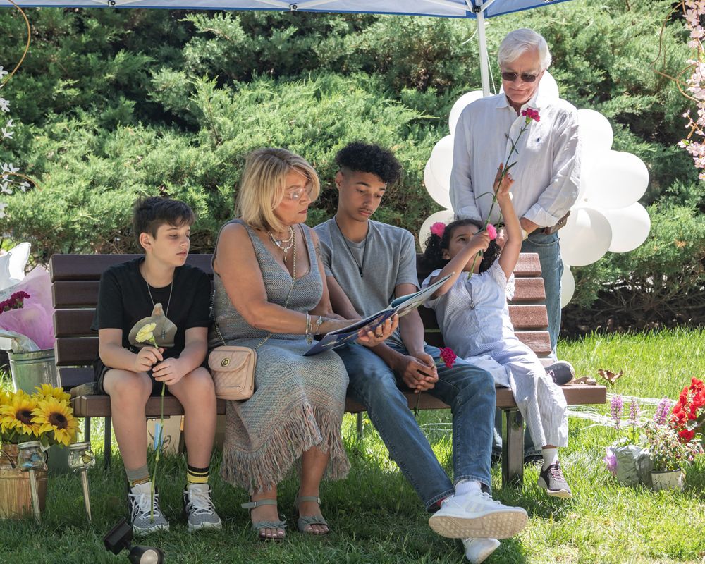 A group sits together on a bench in a garden surrounded by flowers and greenery.