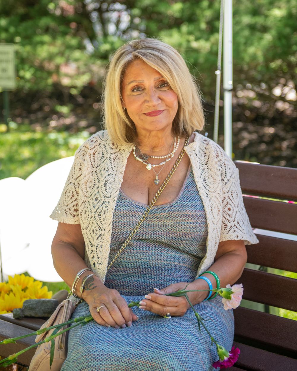Woman in lace cardigan sitting on outdoor bench with flowers and accessories.