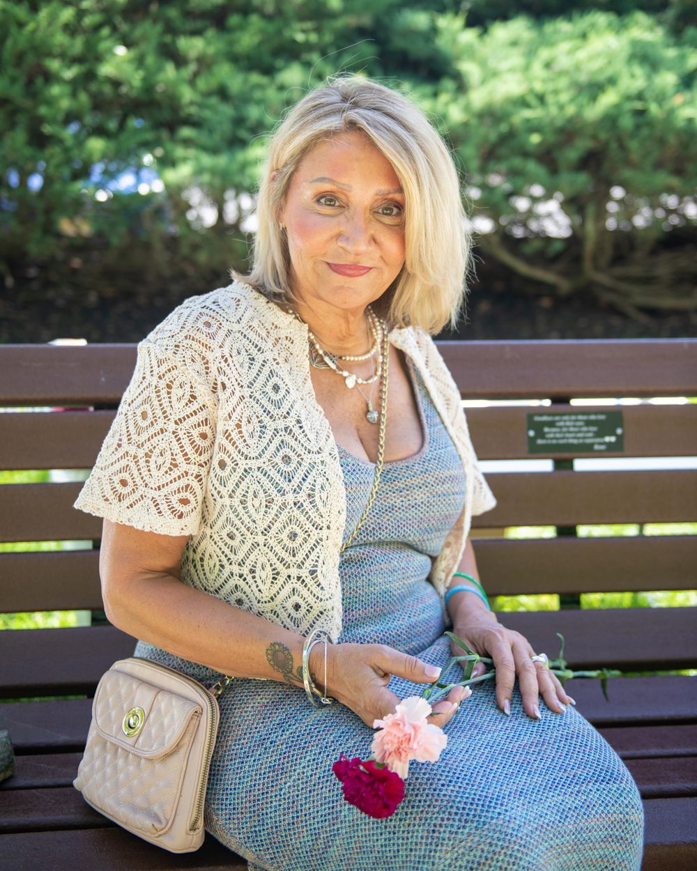 A person sits on a wooden bench wearing a patterned vest and blue dress while holding pink flowers.