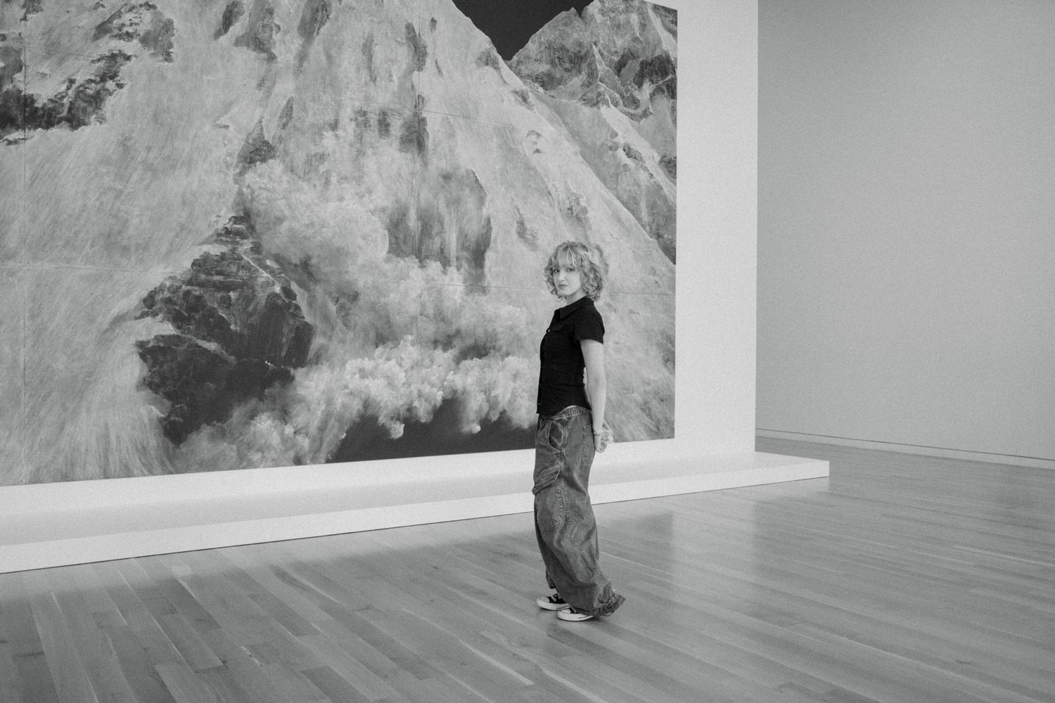Person in black outfit standing before large mountain artwork in minimalist gallery space.