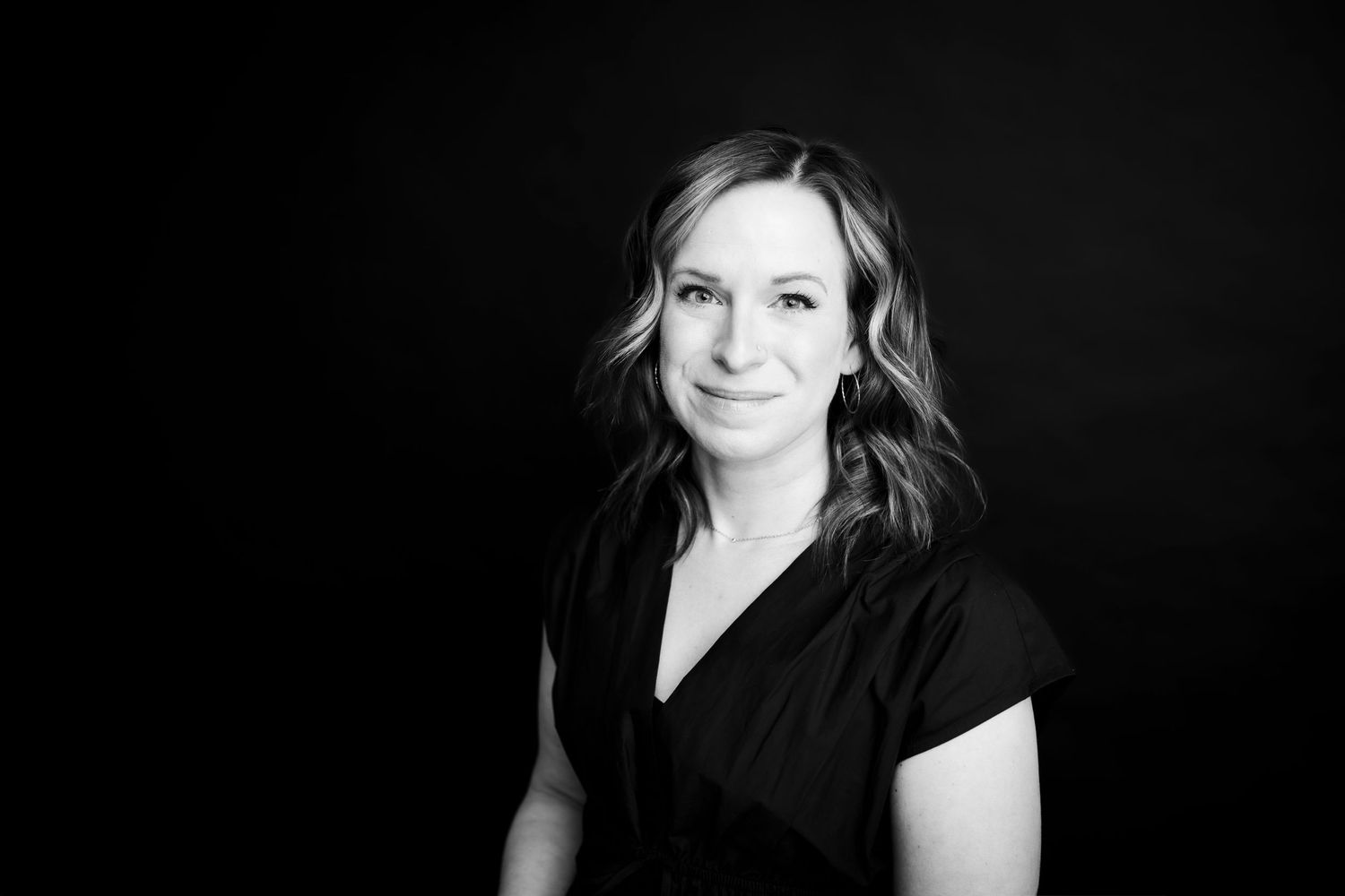 Black and white professional portrait of a person in a black top smiling warmly against a dark background.