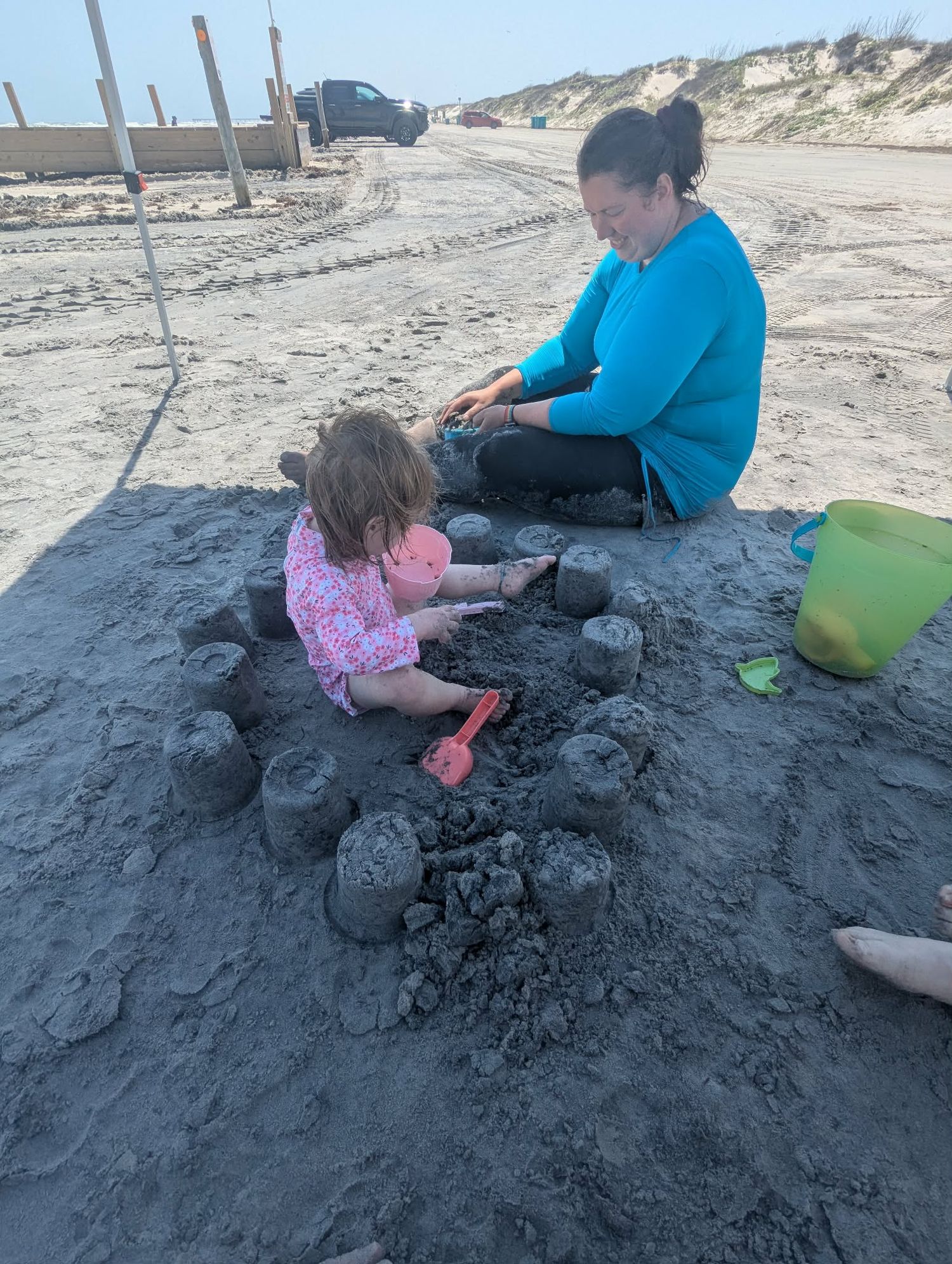 A woman and toddler girl build sand castles together on a beach using colorful plastic buckets and shovels.