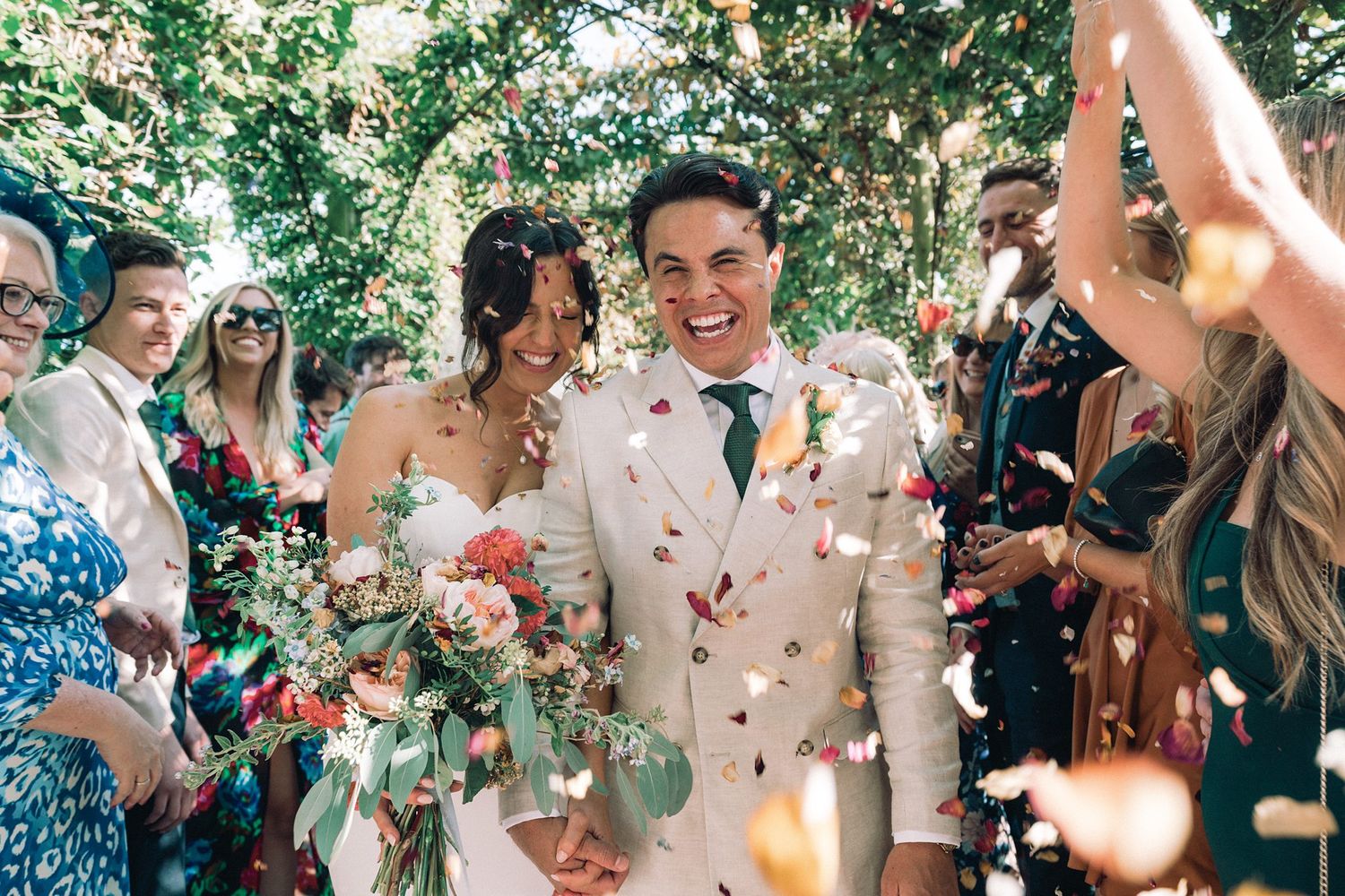 Joyful newlyweds walk through confetti shower surrounded by smiling guests at outdoor garden wedding ceremony.