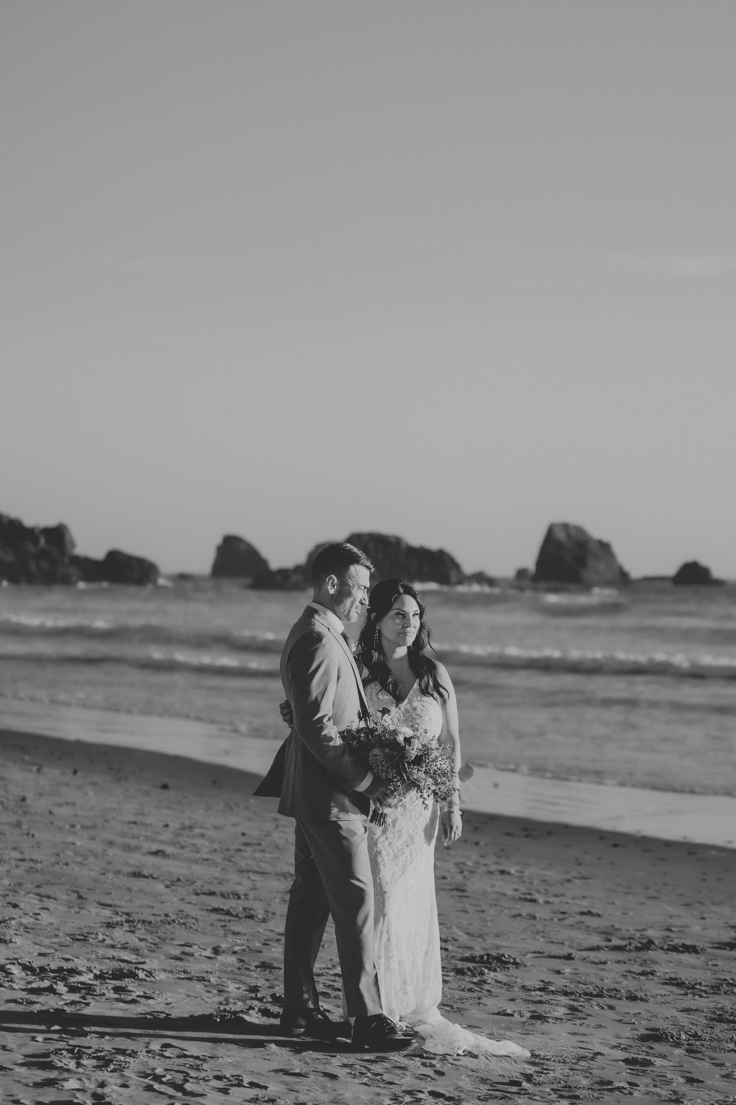 Black and white photo of a bride and groom embracing on a sandy beach with ocean waves and rocks in the background.