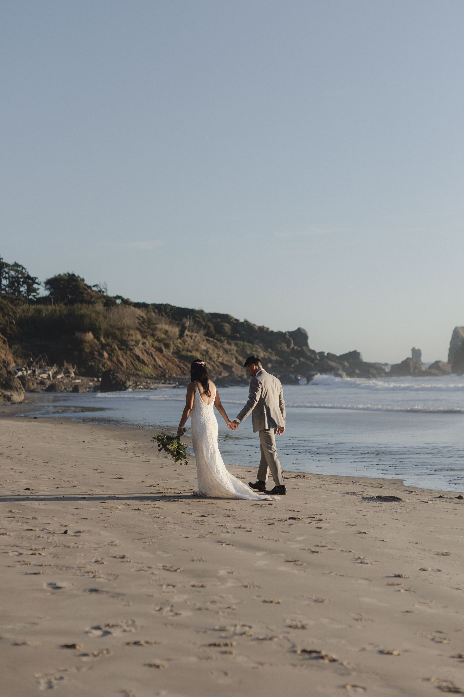 Bride and groom hold hands walking along a sandy beach shoreline, bride in white gown holding bouquet.