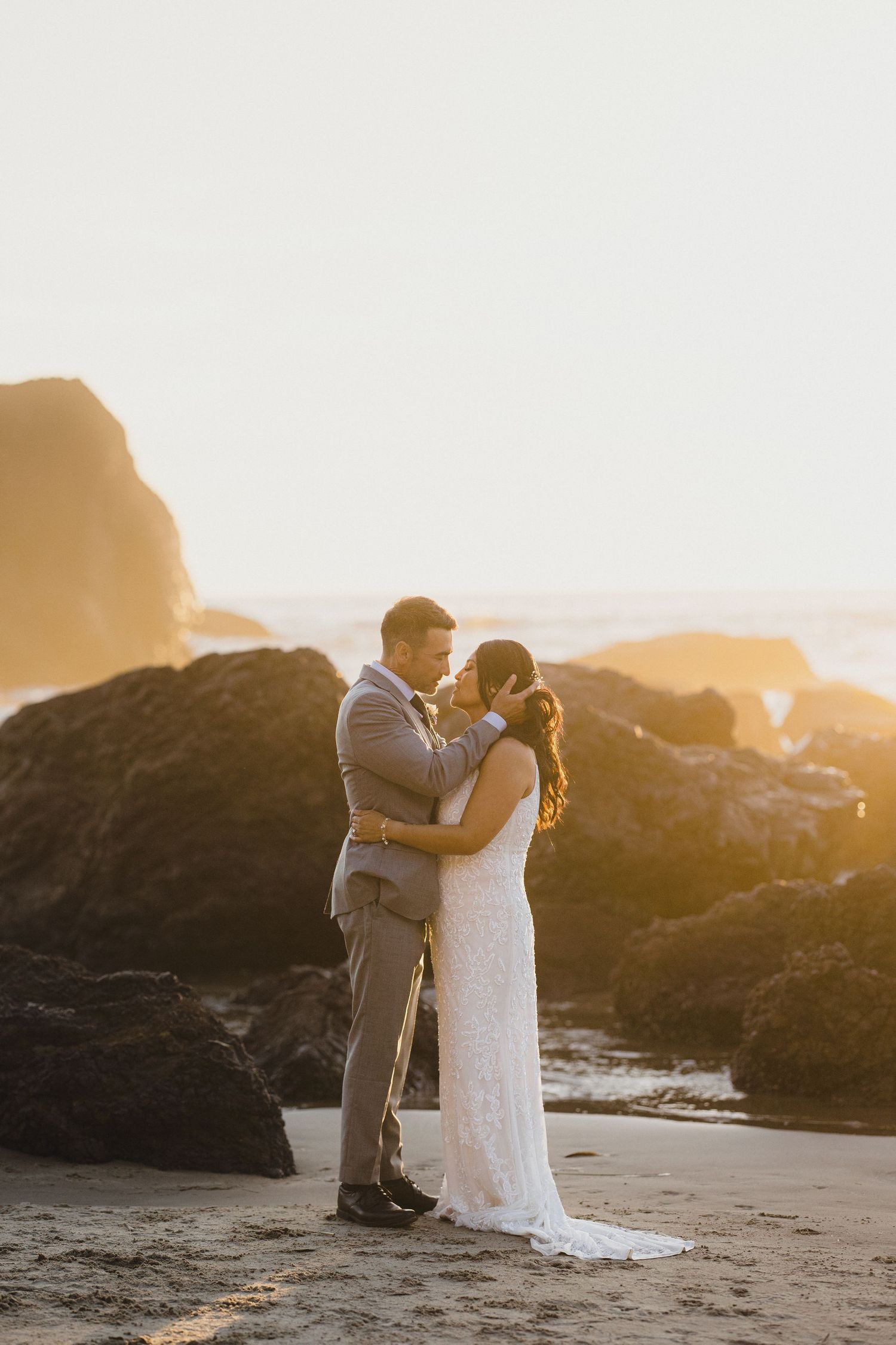 Bride and groom embrace on a sandy beach at golden sunset, with dramatic rocky cliffs in the background.
