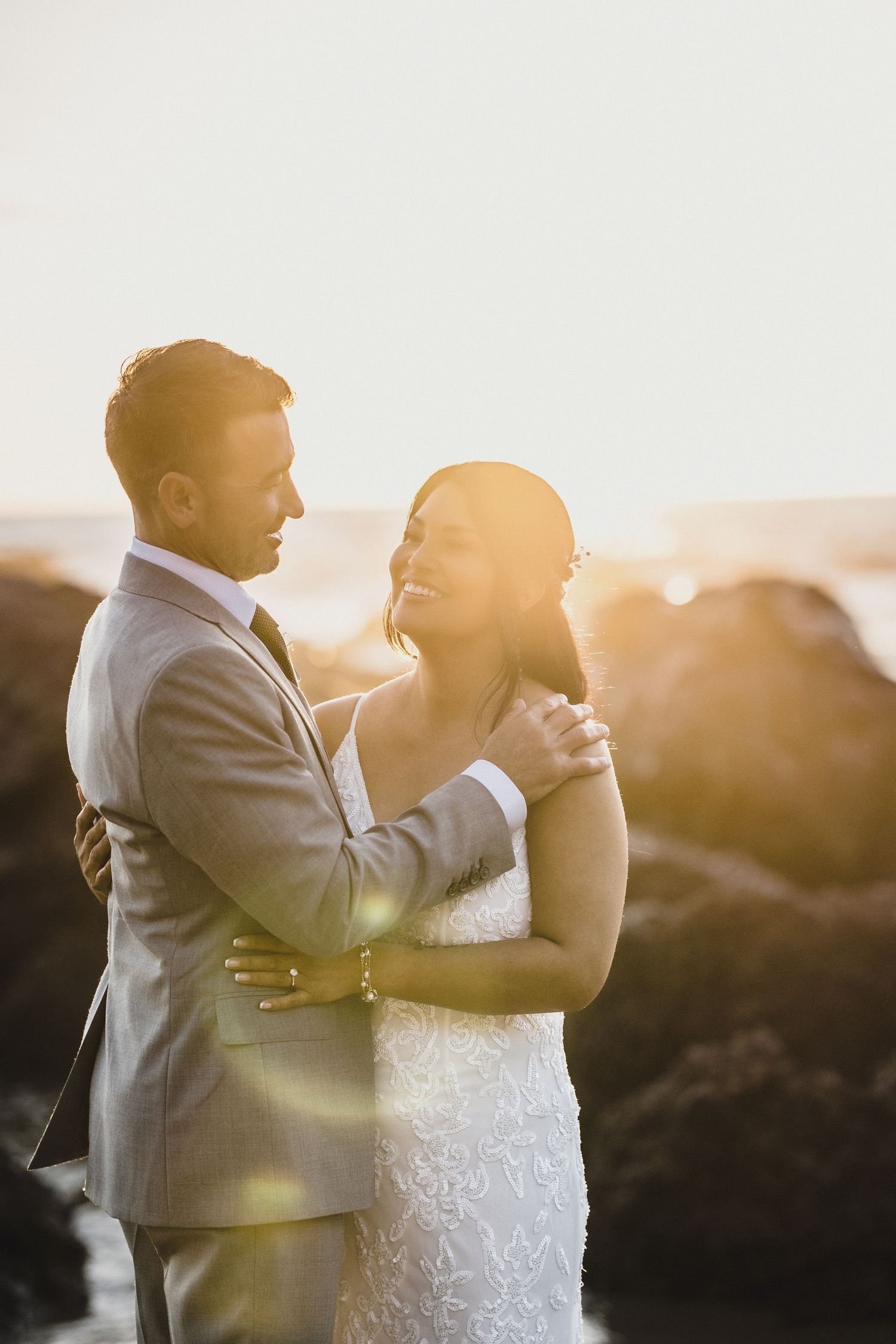 Couple embraces at golden sunset on rocky beach, groom in gray suit holding smiling bride in lace gown.