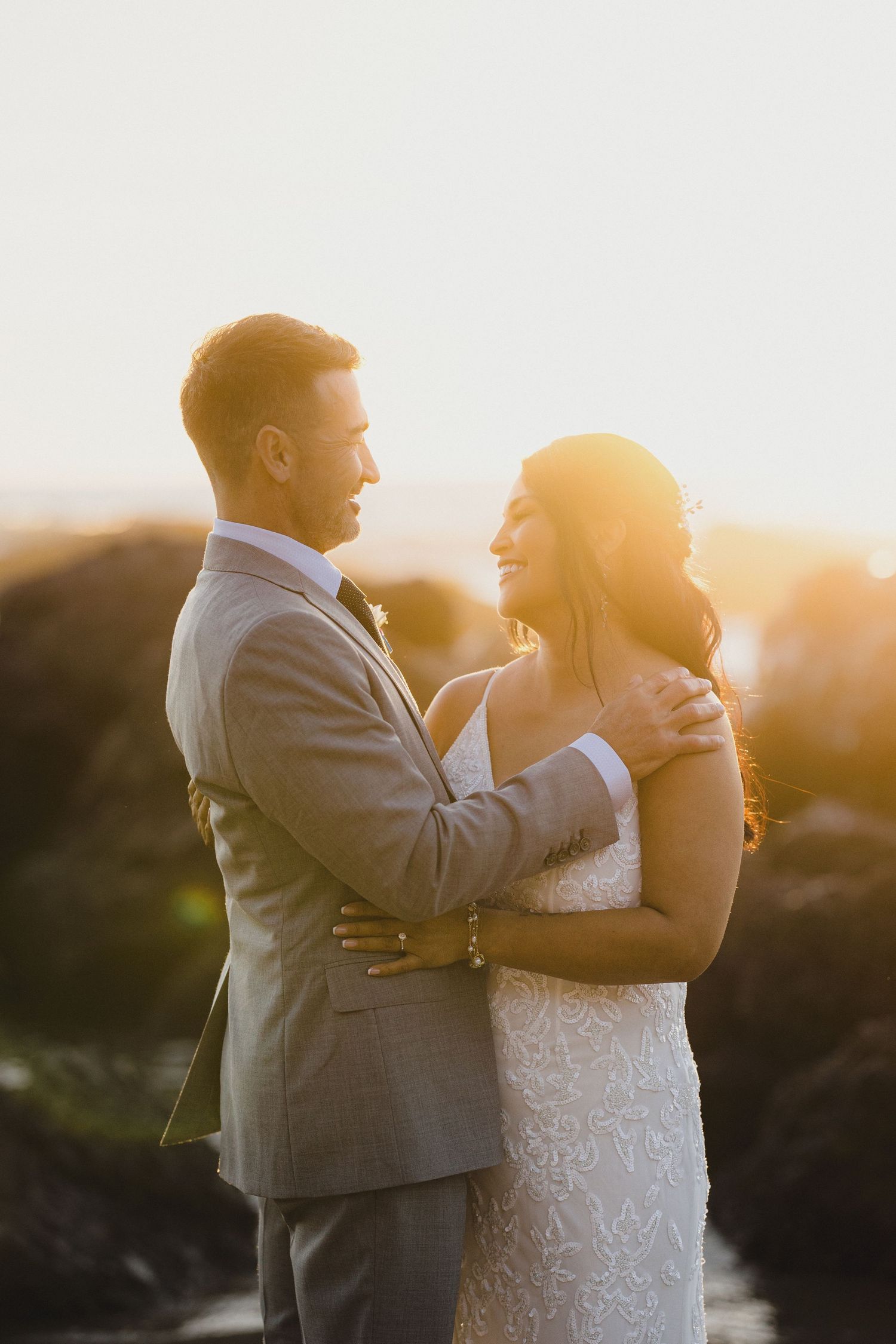 A bride and groom embrace during a golden sunset, the groom in a gray suit and bride in a lace wedding gown.