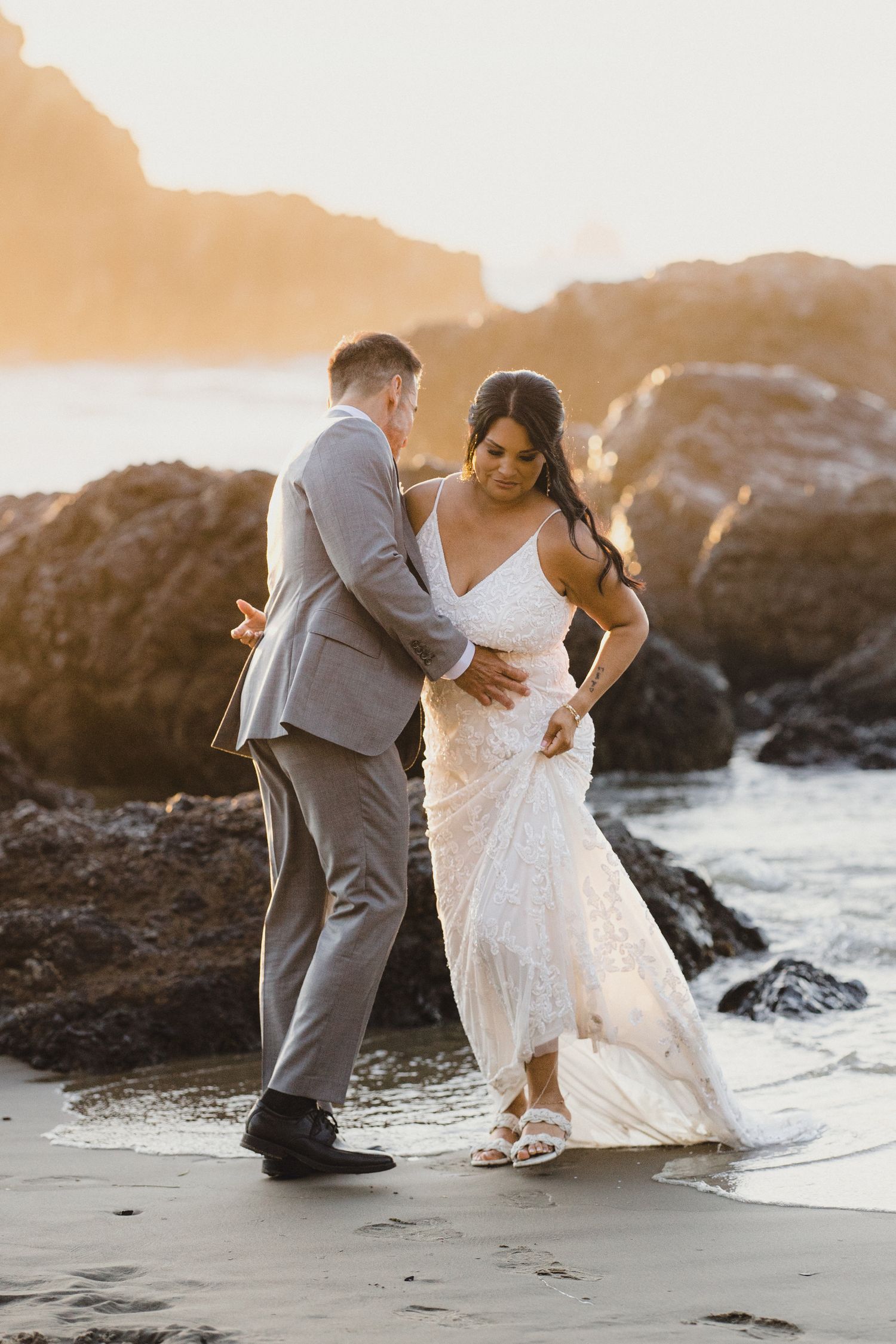 Groom in gray suit helps bride in lace wedding dress navigate ocean waves on a rocky sunlit beach.