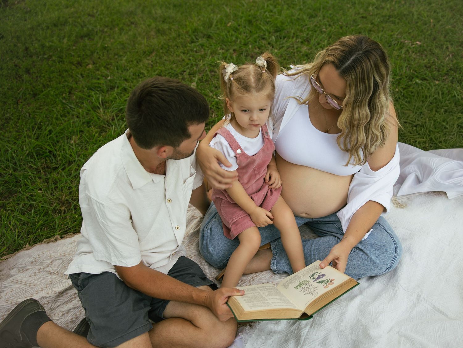 Pregnant woman and man reading a book with toddler girl on a blanket in a green park.
