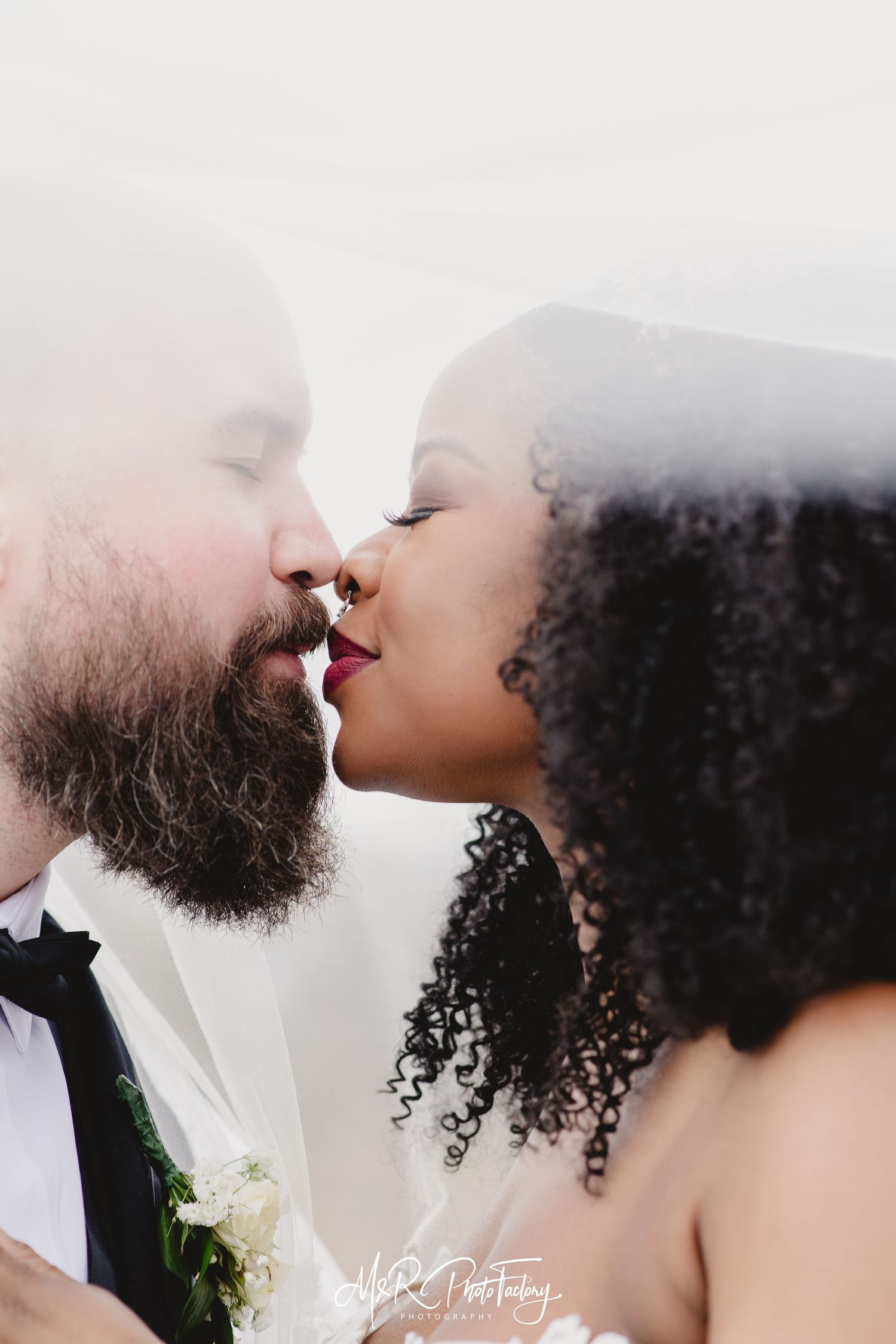 Interracial couple shares a tender kiss under a bridal veil in a close-up wedding portrait.