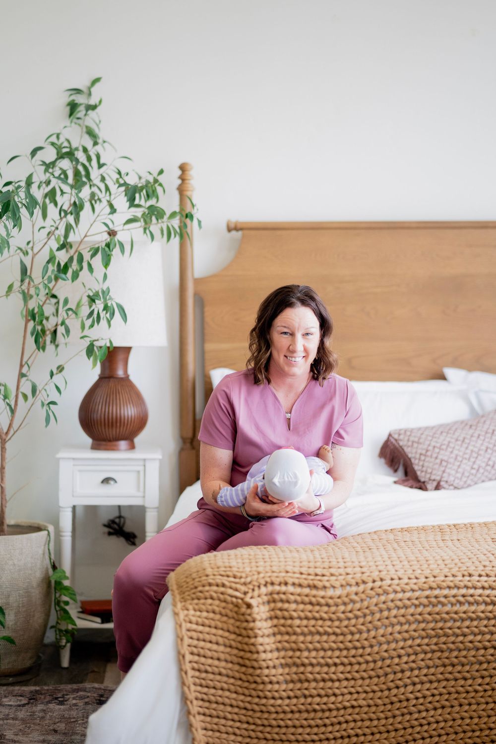 Smiling nurse in pink scrubs sitting on bed holding newborn baby in cozy bedroom setting.